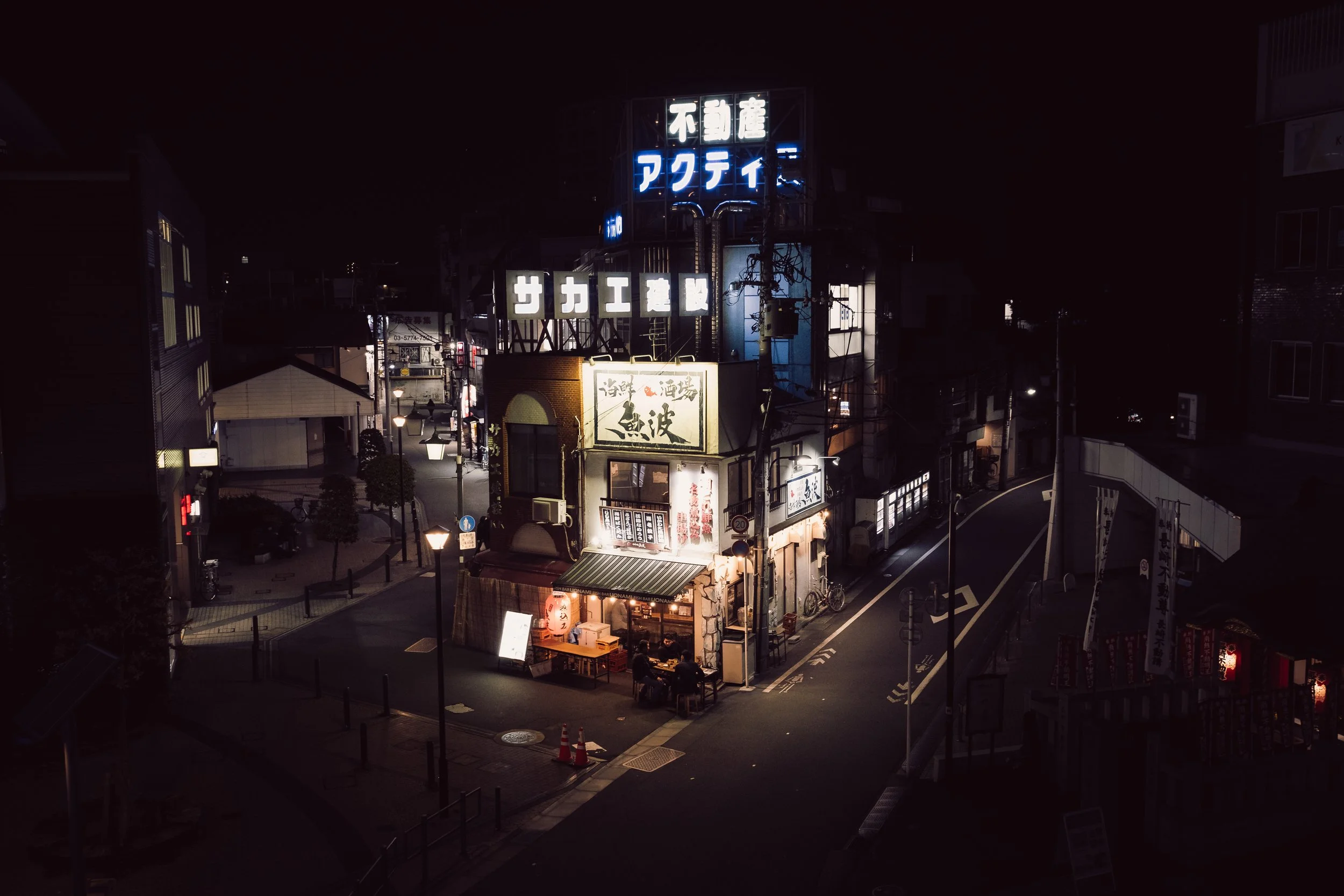 Nighttime street scene in Japan with illuminated signs on a small building and surrounding shops.