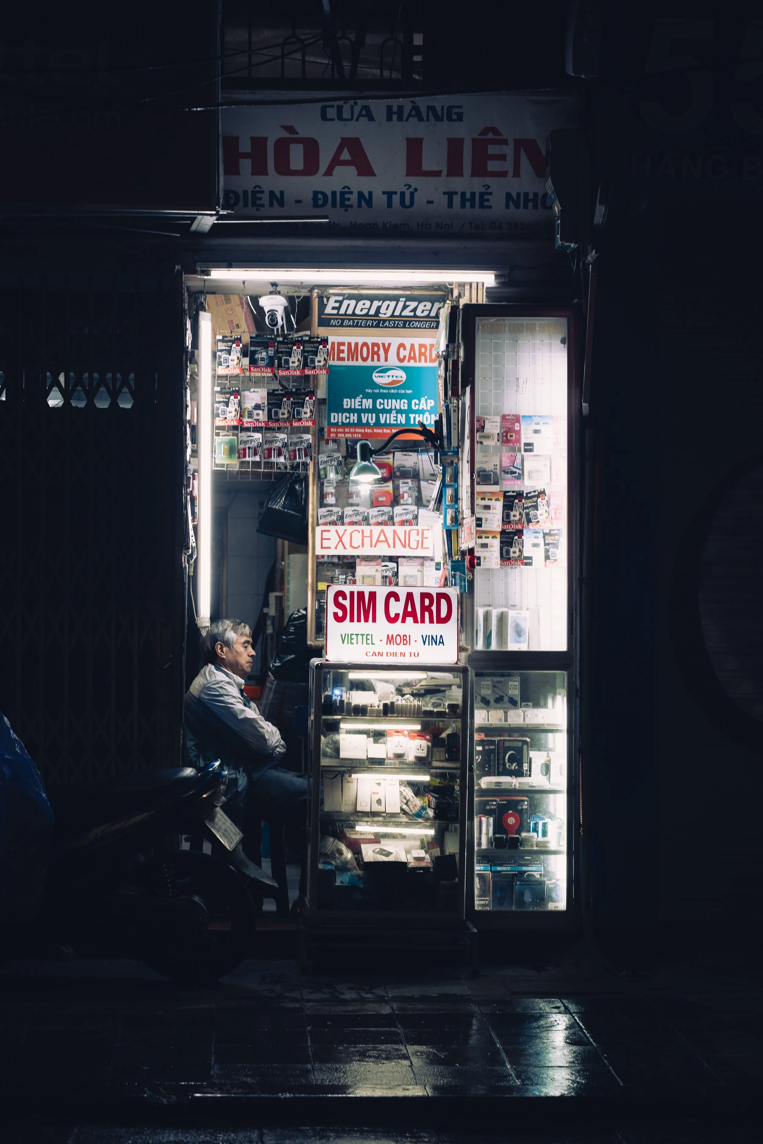 A small electronics shop at night with various items on display, including SIM cards, memory cards, and batteries. An elderly man is sitting inside the shop. The shop is lit up, contrasting with the dark outside environment.