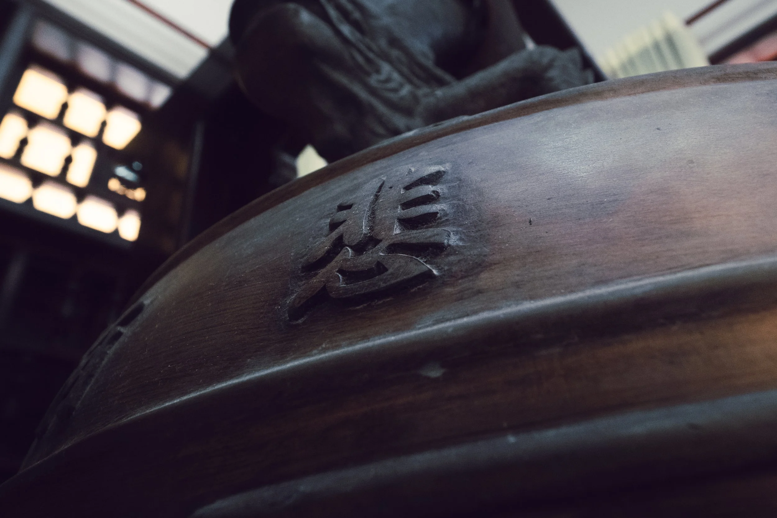 Close-up of a wooden surface with a carved Chinese character in the center, with background elements of a patterned wall and a figure wearing dark clothing.