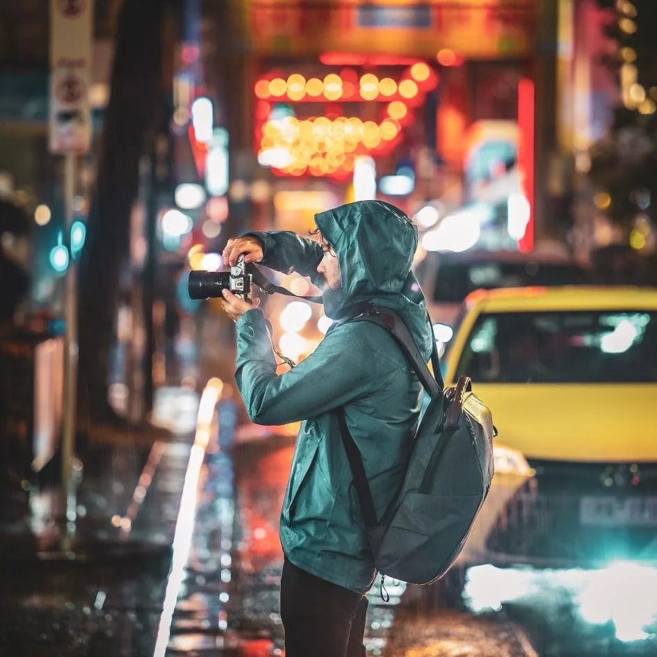 A person in a raincoat takes a photo on a city street at night with colorful blurred lights and cars.