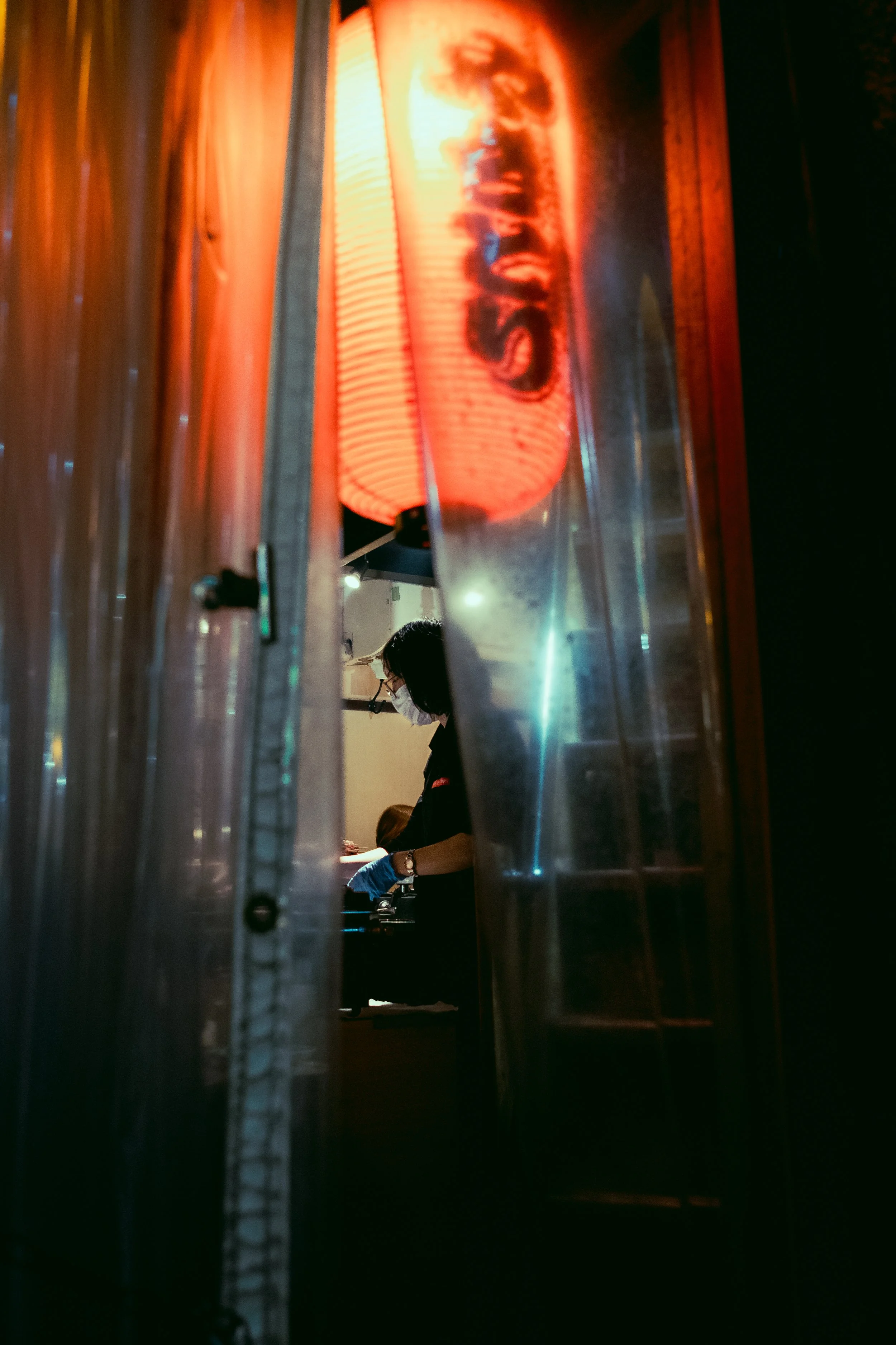 A person wearing a face mask and gloves working inside a restaurant kitchen, viewed through a narrow gap between curtains, with a bright red lantern displaying the word 'SASHI'.