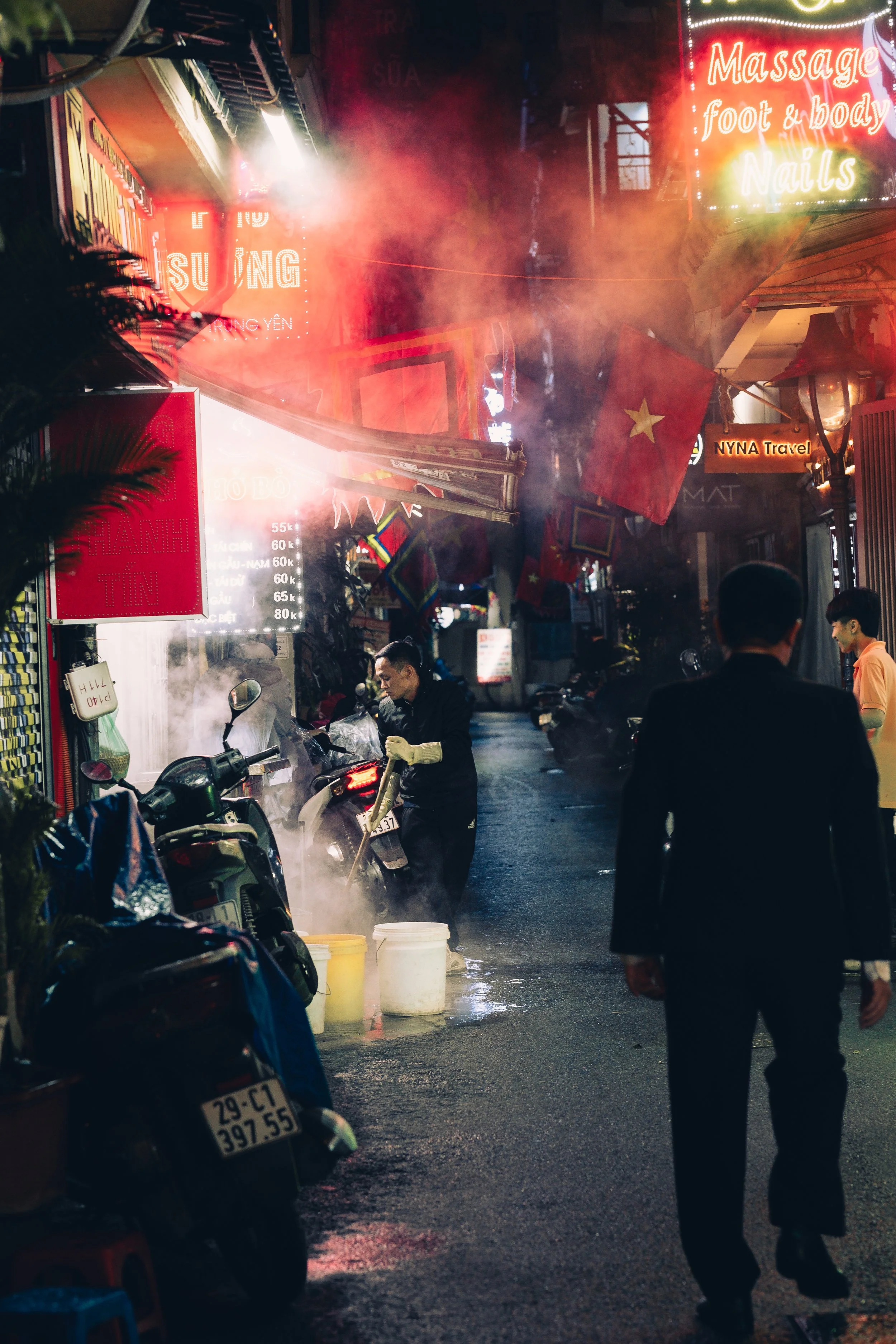 A street scene at night with a person in black clothes and gloves washing a motorcycle, surrounded by parked motorcycles and storefronts with neon signs and flags. Steam rises from the street, and there are people walking and talking.