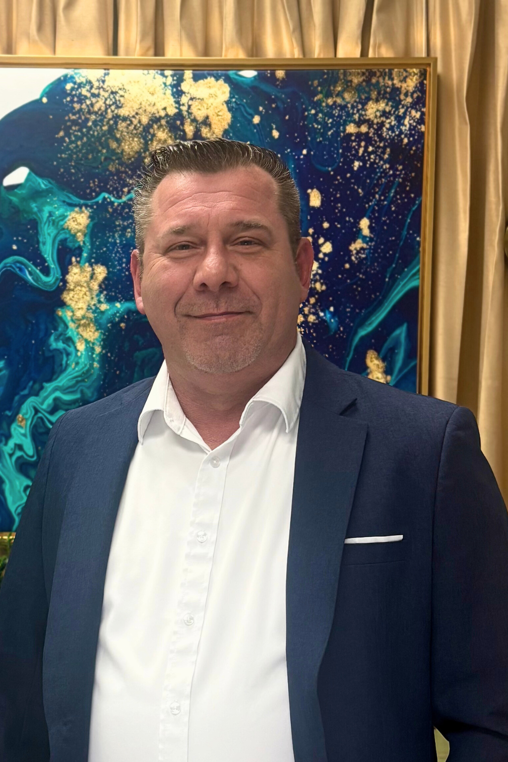 Portrait of John Doyle  in a white dress shirt and navy blazer standing indoors in front of abstract blue and gold artwork with curtains.