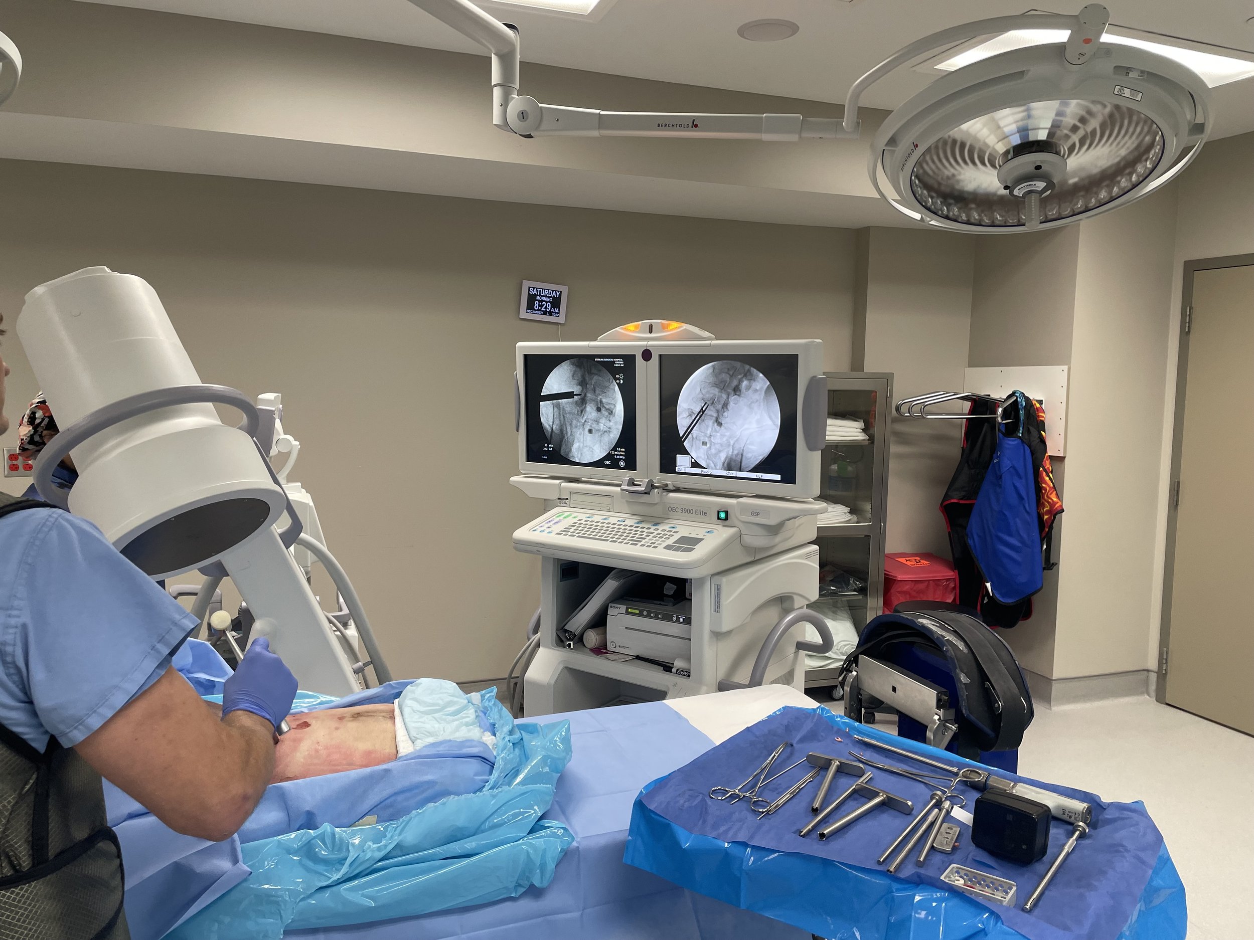 Medical procedure room with a healthcare worker preparing a patient for a procedure, with monitors displaying X-ray images and surgical tools on a blue sterile drape.