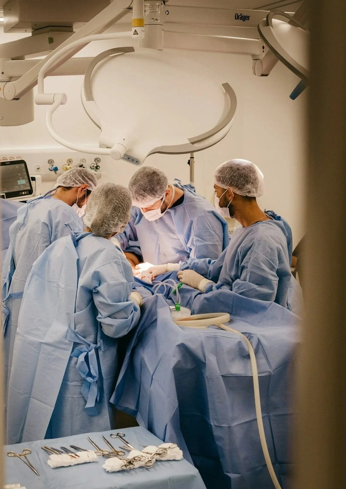 A team of surgeons dressed in sterile blue scrubs, surgical masks, and hair covers working together during an operation in an operating room, with surgical tools placed on a tray nearby.