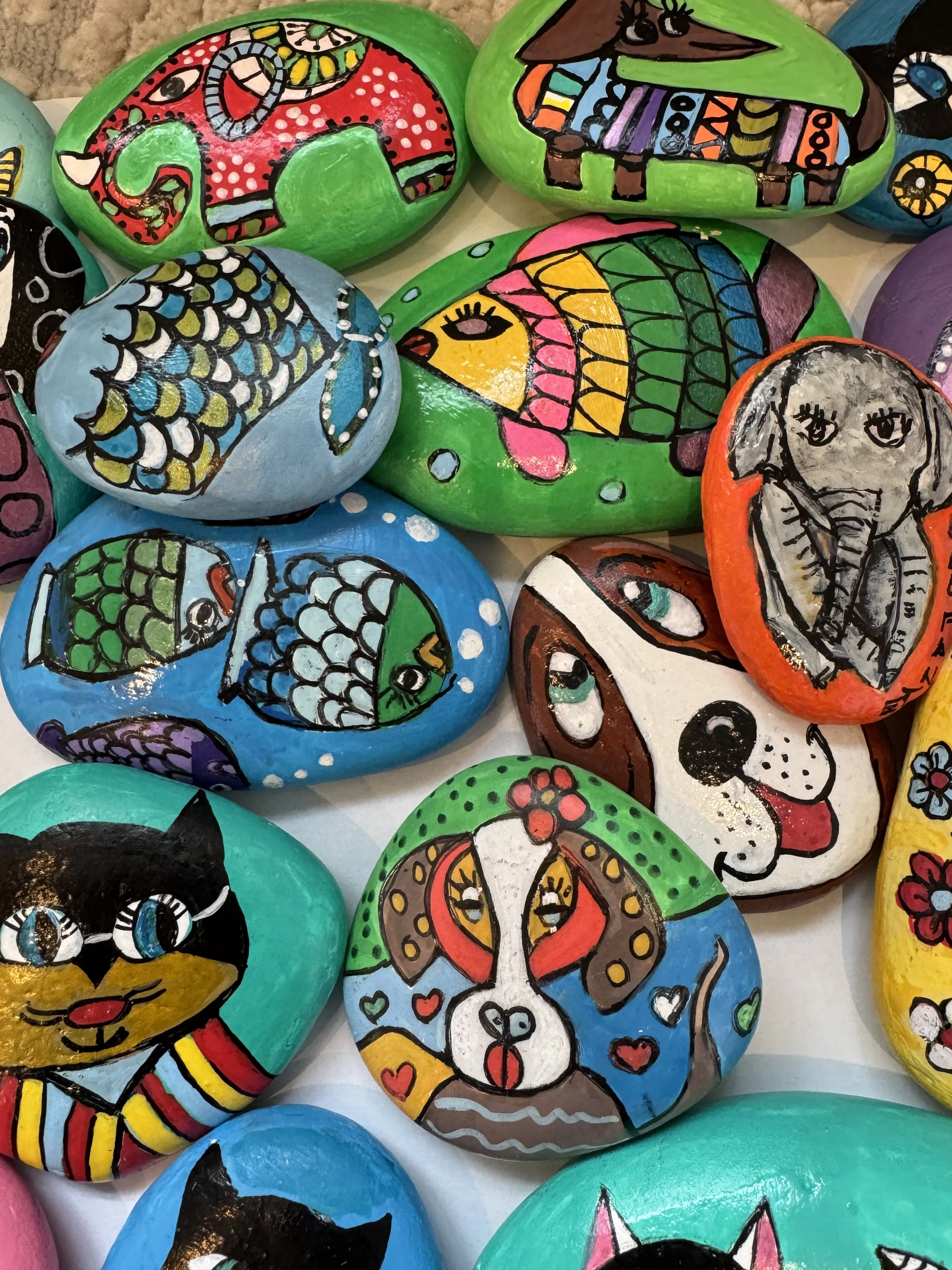 Colorful painted rocks featuring various animals, including a cat, dog, elephant, fish, and a pig, with decorative patterns and designs.
