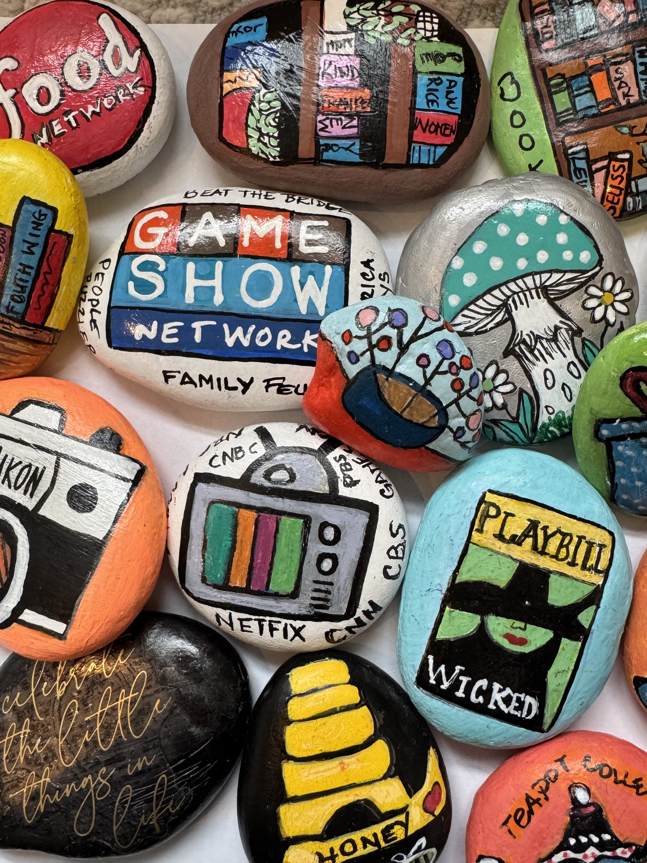Colorful painted rocks with various designs and messages, including a book shelf, game show, mushroom, camera, Netflix screen, Playbill, and honey pot.
