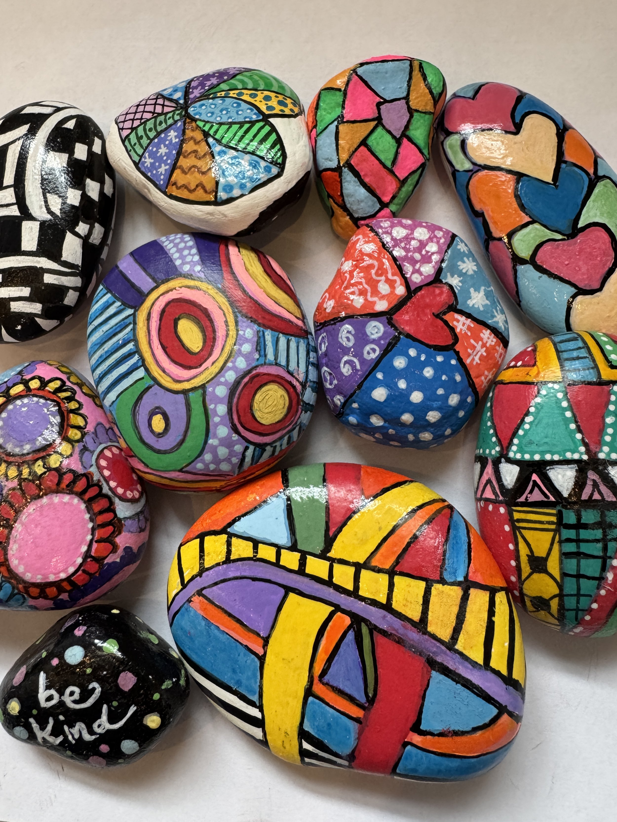 Colorful painted rocks with various patterns and designs, including hearts, flowers, and abstract shapes, arranged on a white surface.