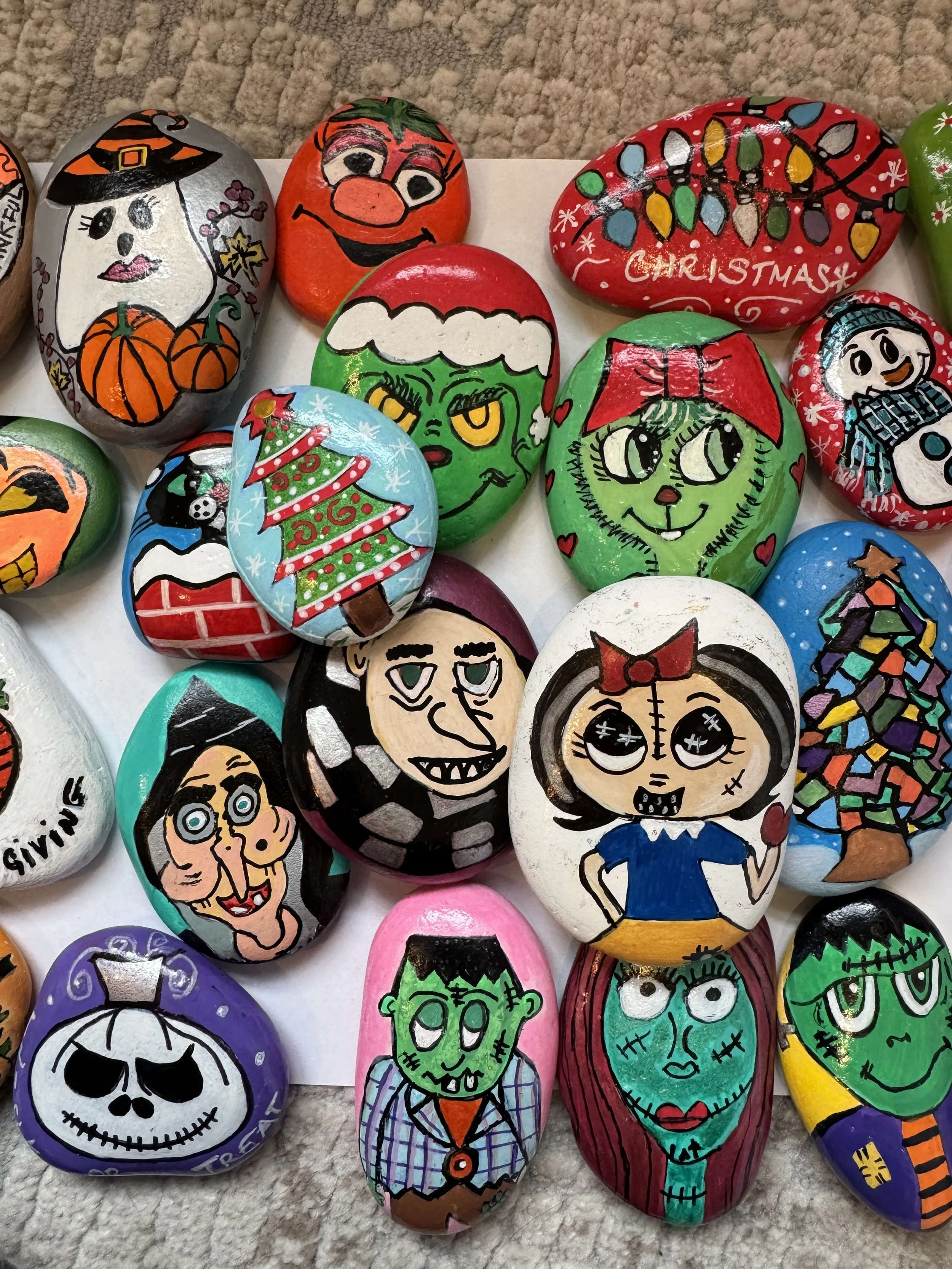 Colorful painted rocks featuring Halloween and Christmas themed characters and designs, including a witch, a pumpkin, Santa, a Christmas tree, a zombie, and a snowman.