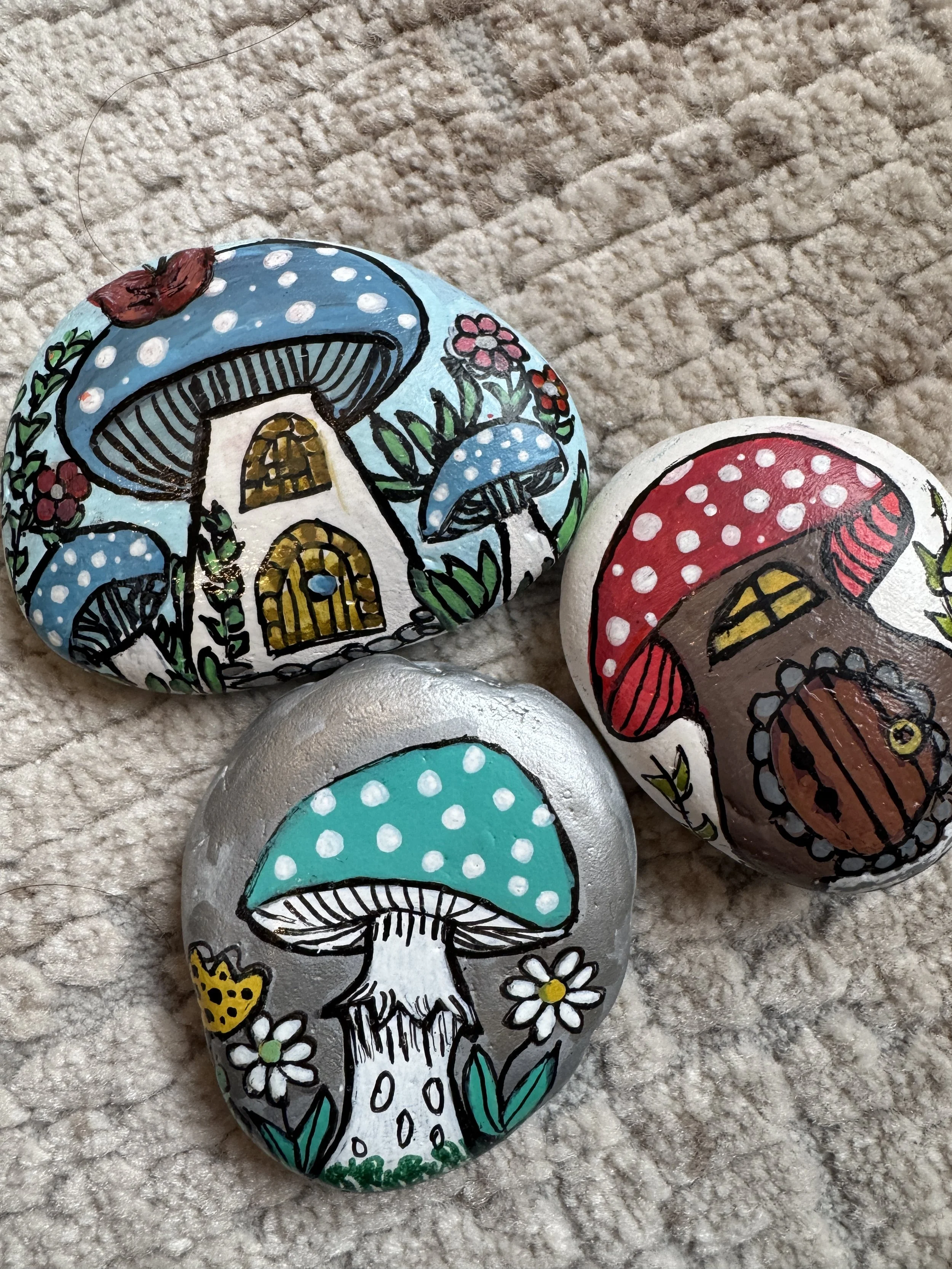 Painted rocks with colorful mushroom designs, including a large blue mushroom with a house underneath, a red mushroom with a cabin underneath, and a teal mushroom with white spots surrounded by flowers.