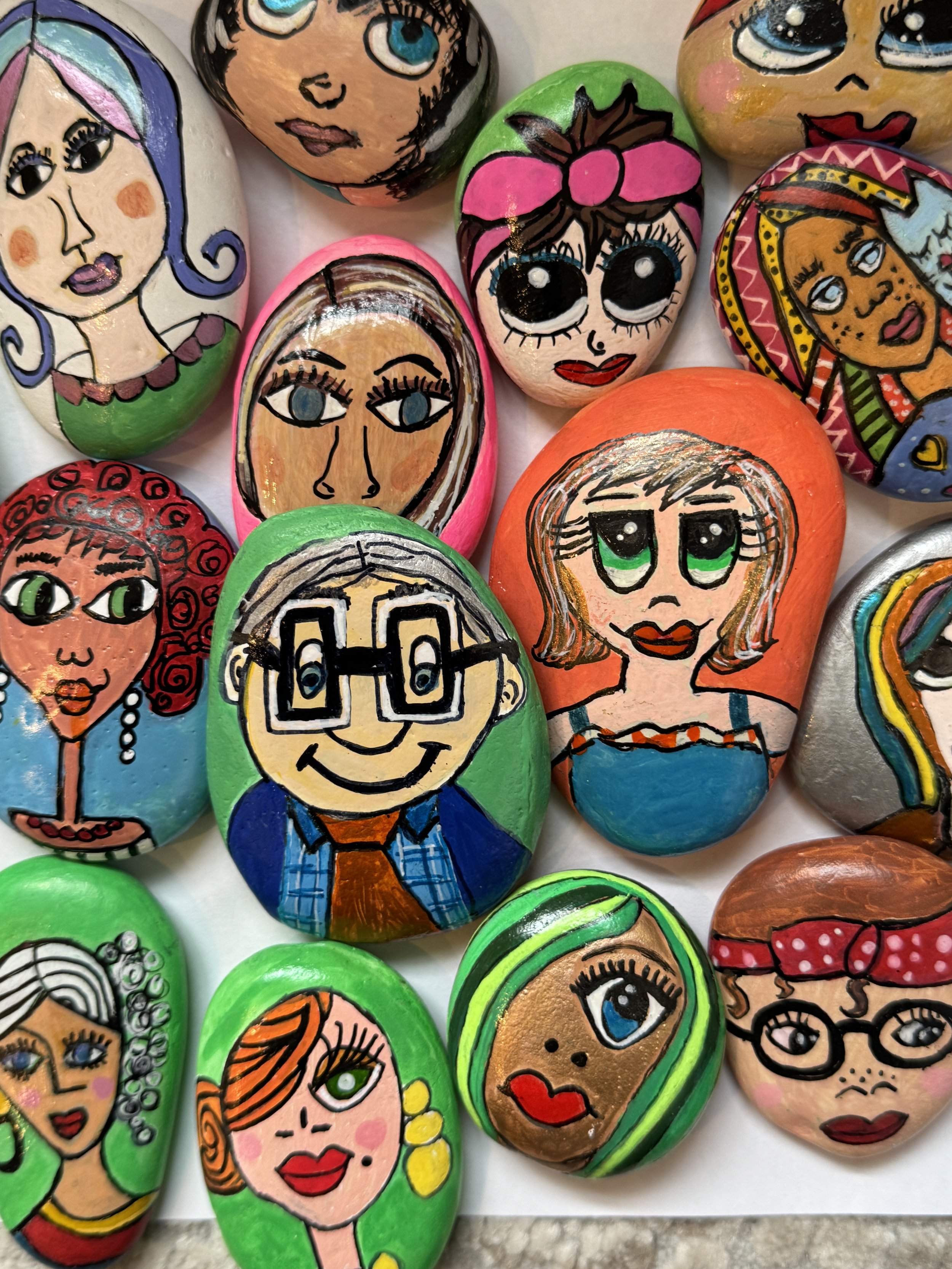 Colorful painted rocks with illustrated faces of women and one man, featuring different hairstyles, accessories, and expressions, arranged on a flat surface.