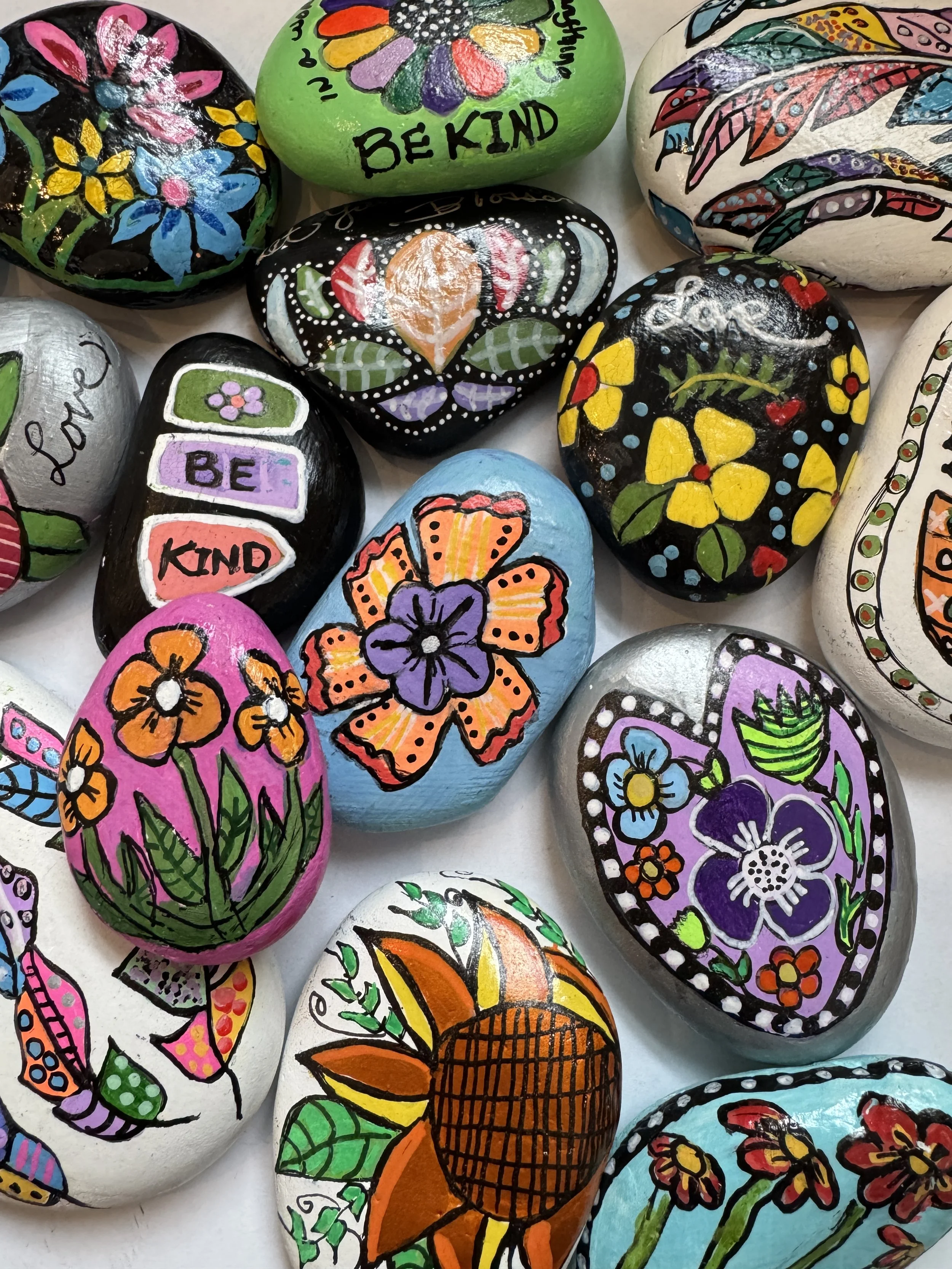 Colorful painted stones with floral and motivational designs, including words like "BE KIND" and various flower illustrations.