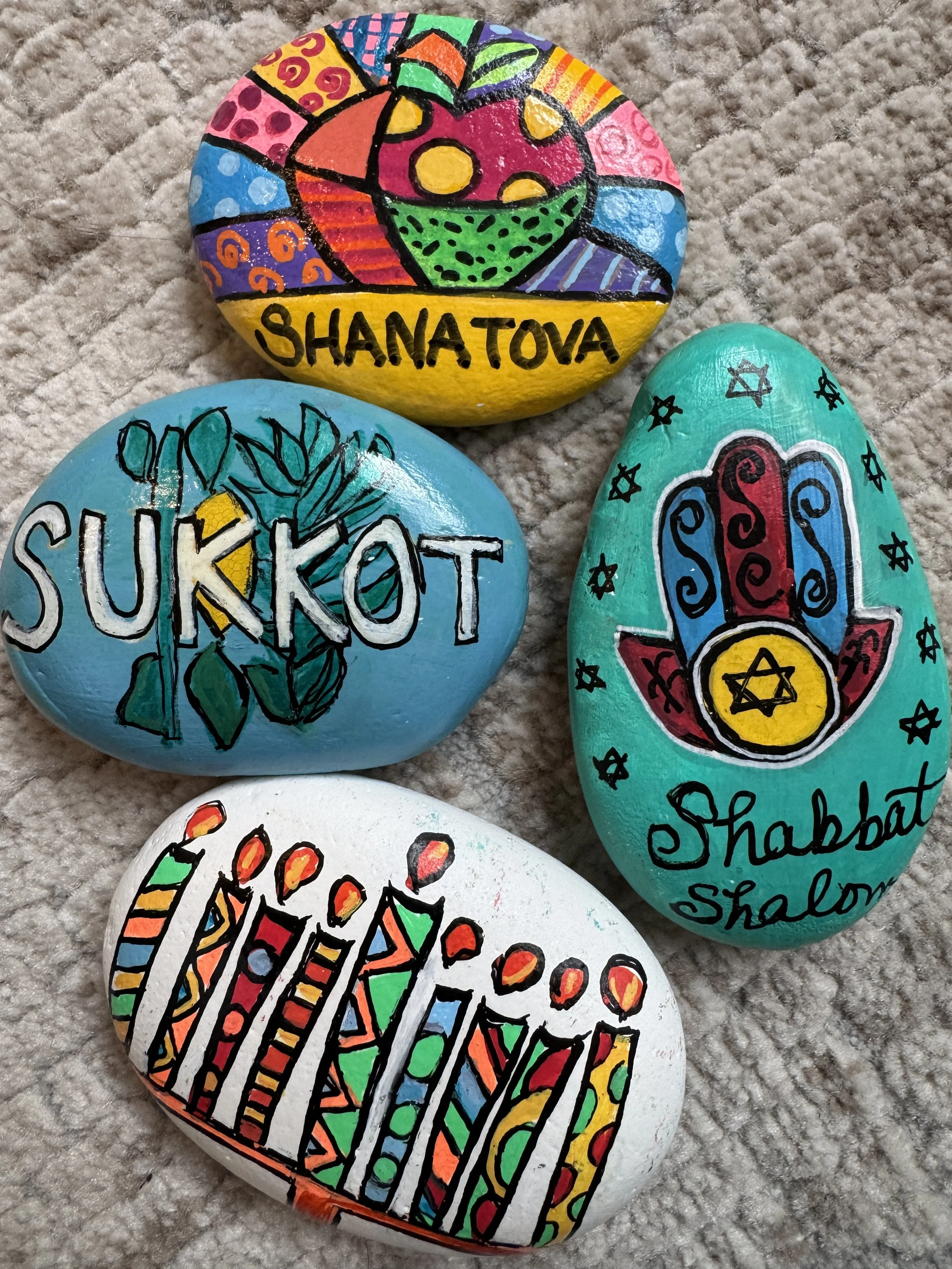Colorfully decorated painted rocks with various symbols and names, including one with a bowl of food and the name 'Shanatova', another with the word 'Sukkot' and a palm branch, a third with a Hamsa hand and the words 'Shabbat Shalom', and a fourth wi