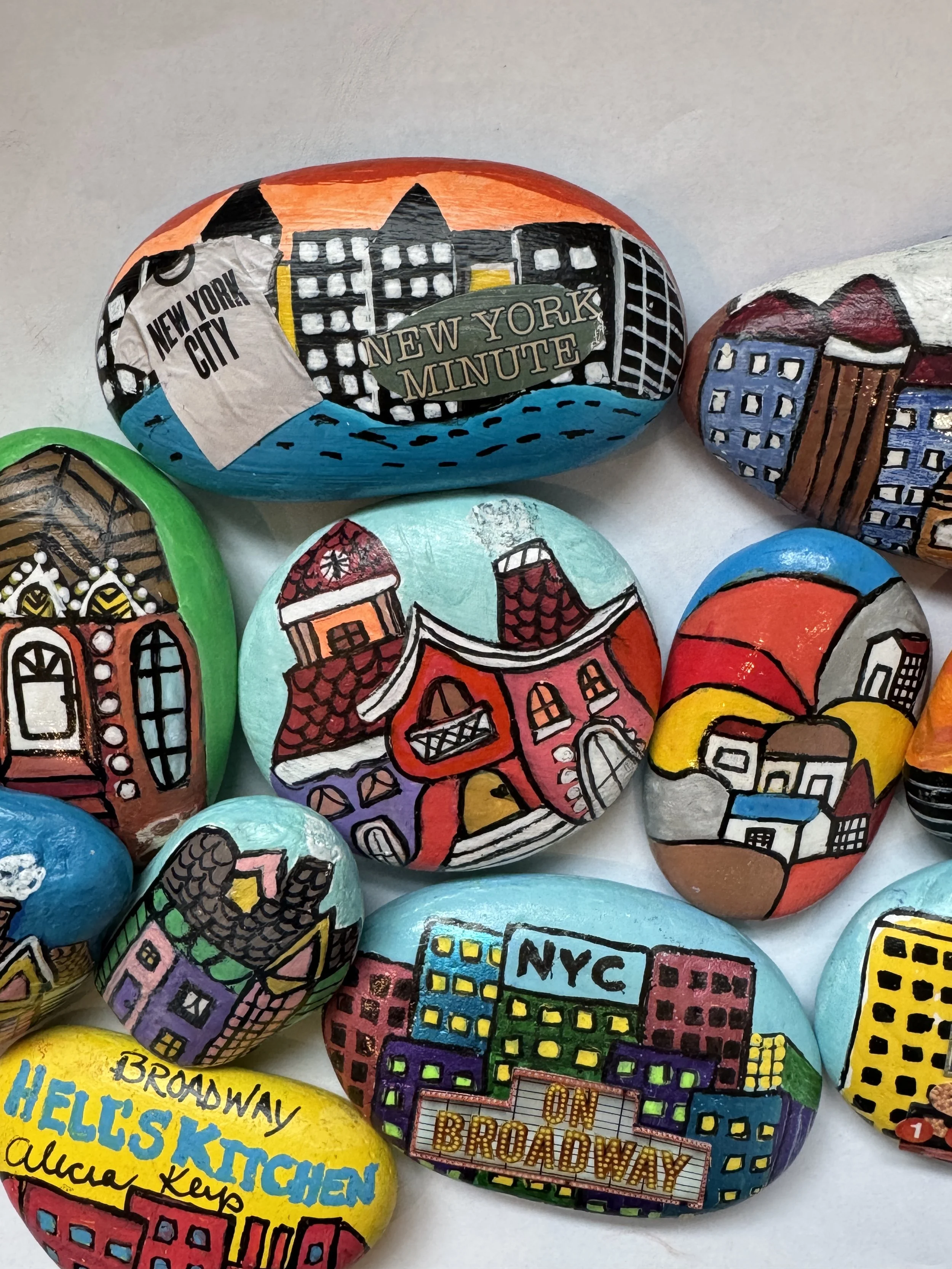 Colorful painted rocks with New York City themed designs, including buildings, landmarks, and signs such as 'New York Minute,' 'NYC on Broadway,' and 'Helskitchen.'