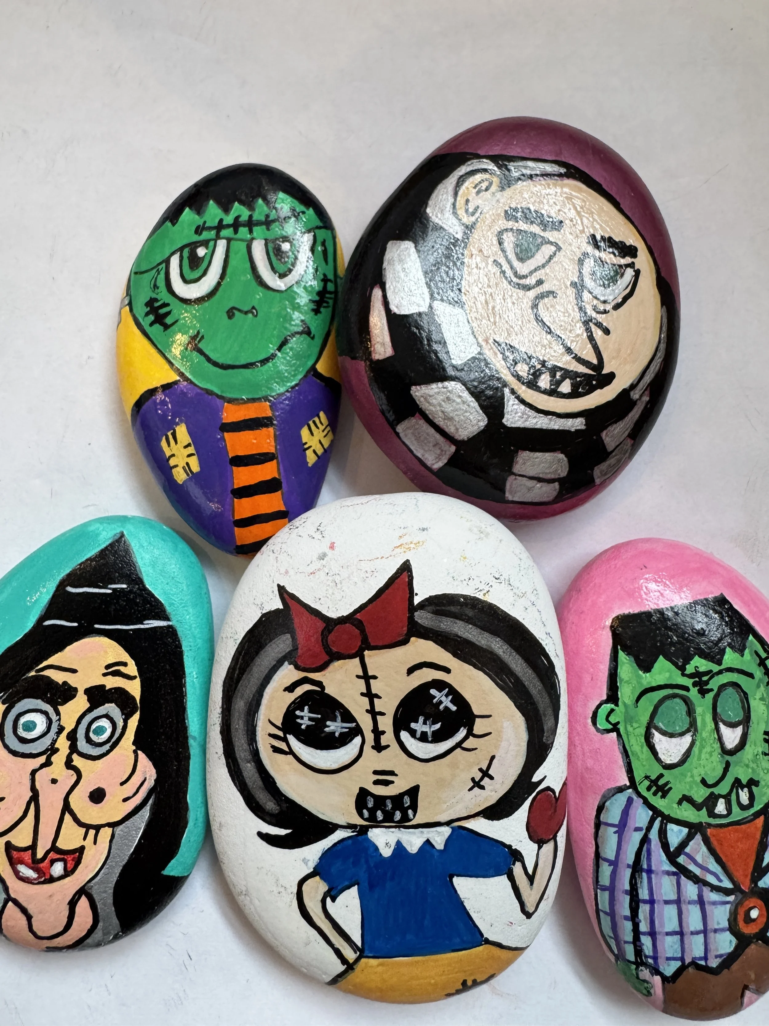 Hand-painted rocks with cartoon character faces, including Frankenstein, a girl with a bow, a witch, a vampire, and a smiling goblin.