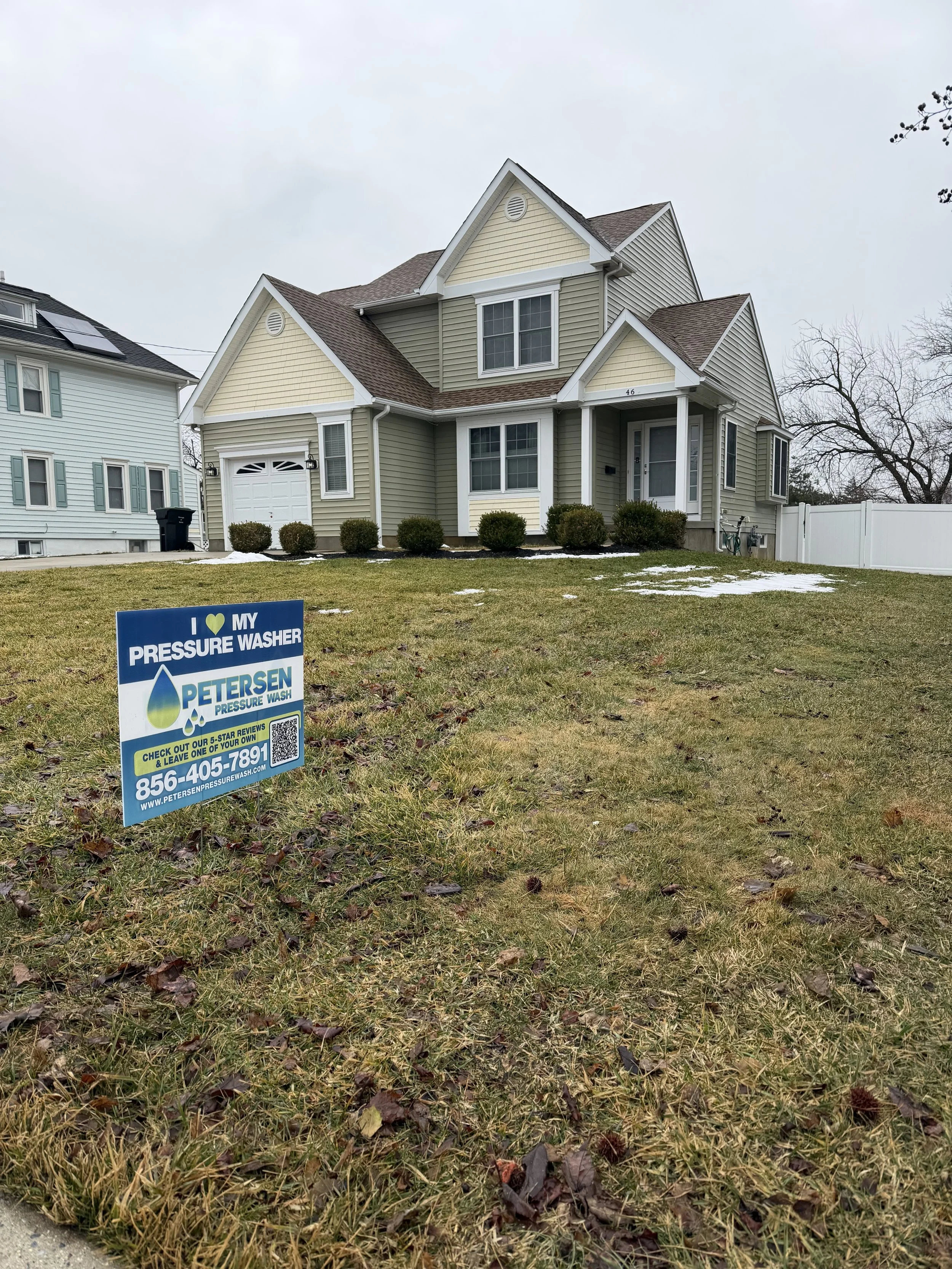 A residential front yard with a house in the background and a sign in the grassy lawn in the foreground. The house is a two-story, yellow with beige siding, with a gabled roof, front porch, and bushes lining the front. The sign advertises Petersen Pressure Wash and includes a phone number and website.