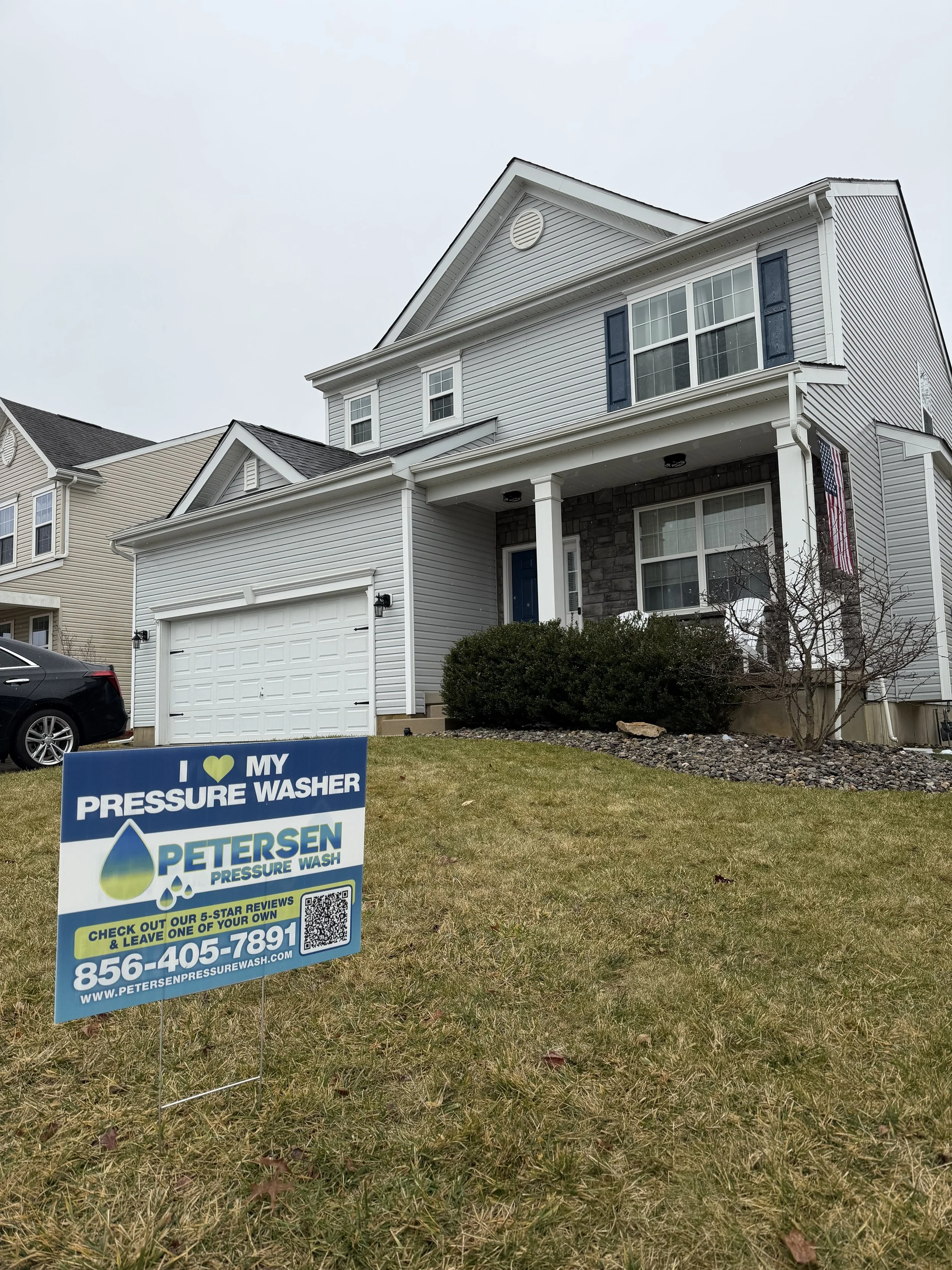 Front yard with a house, a lawn sign for Petersen Pressure Wash, a tree, and neighboring houses visible in the background.