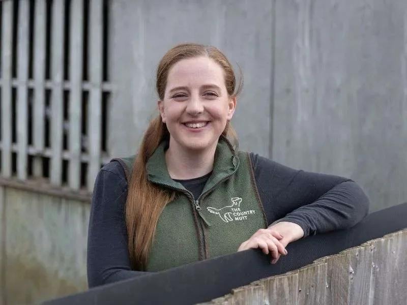 Emily, Dog Handler at The Country Mutt. A woman with long red hair smiling, wearing a Country Mutt green vest with a logo on it, standing outdoors beside a wooden fence.