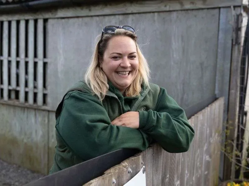 Mille Hodge, owner of The Country Mutt. A smiling woman with blonde hair, wearing a 'The Country Mutt' branded green jacket and sunglasses on her head, leans on a wooden fence outdoors.