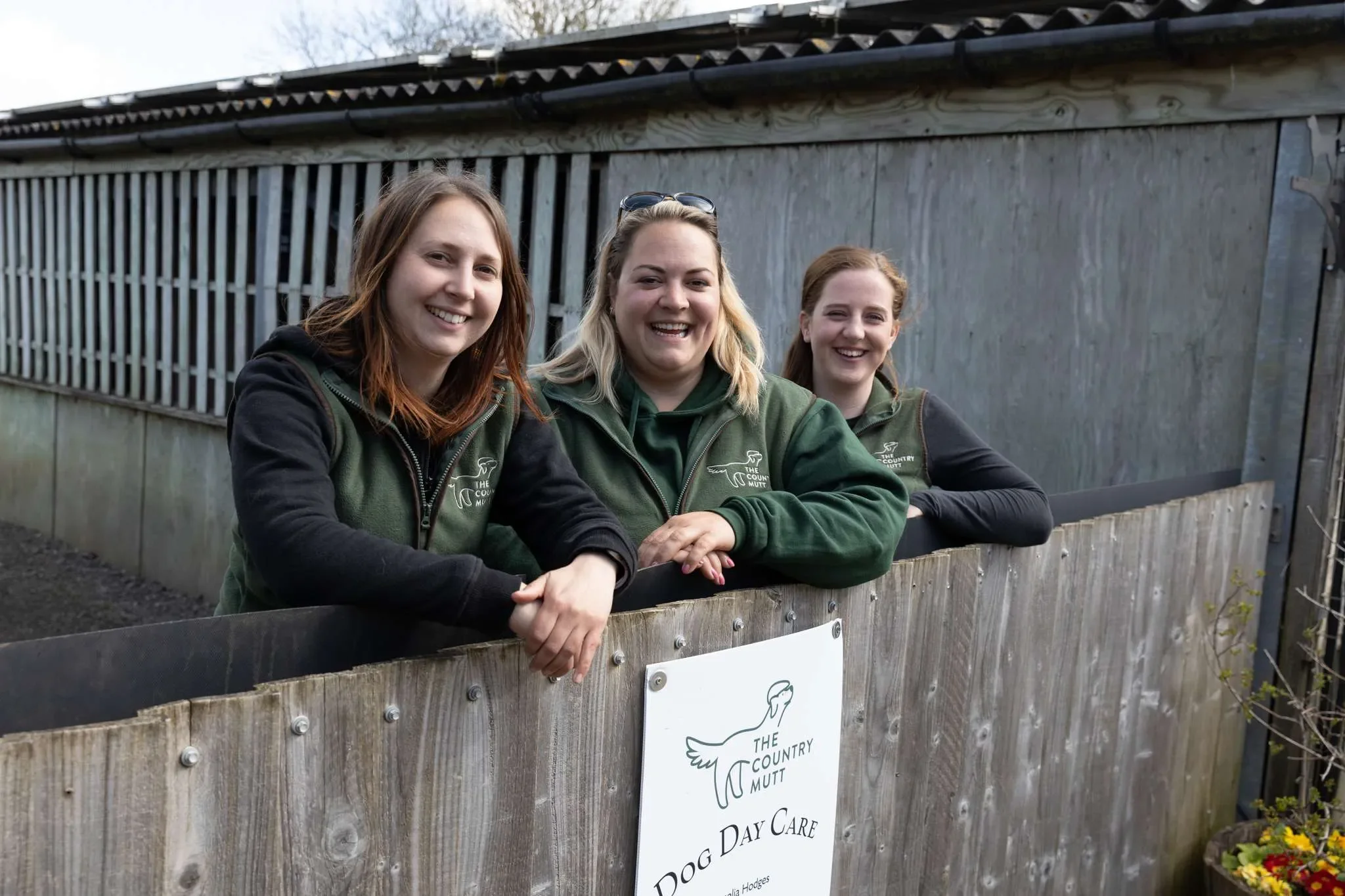 Three women standing behind a wooden fence at a dog day care, smiling at the camera. They are wearing green jackets with a logo that says 'The Country Mutt.' A sign on the fence reads 'Dog Day Care' and features a drawing of a dog.