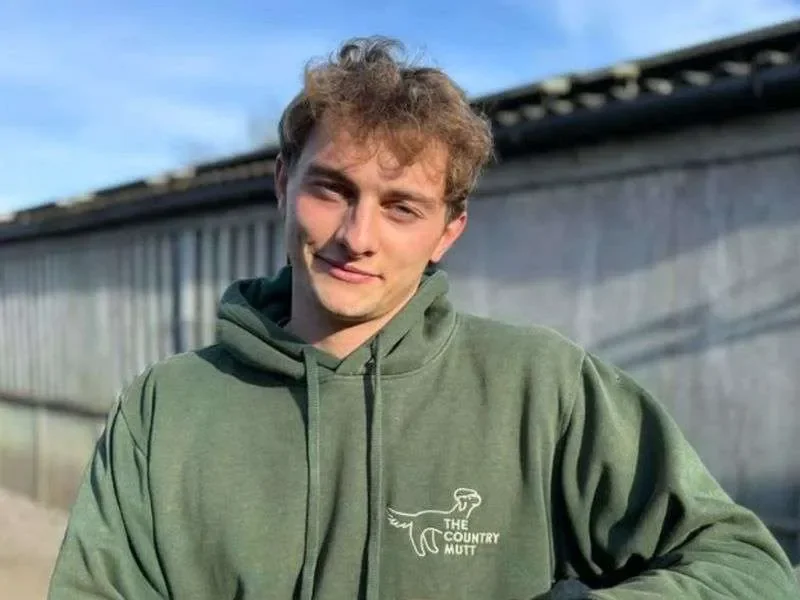 Pete, Dog Handler at The Country Mutt. Young man with curly brown hair wearing a green hoodie with a logo that says 'The Country Mutt' and a dog illustration, standing outdoors in front of a building under a cloudy sky.