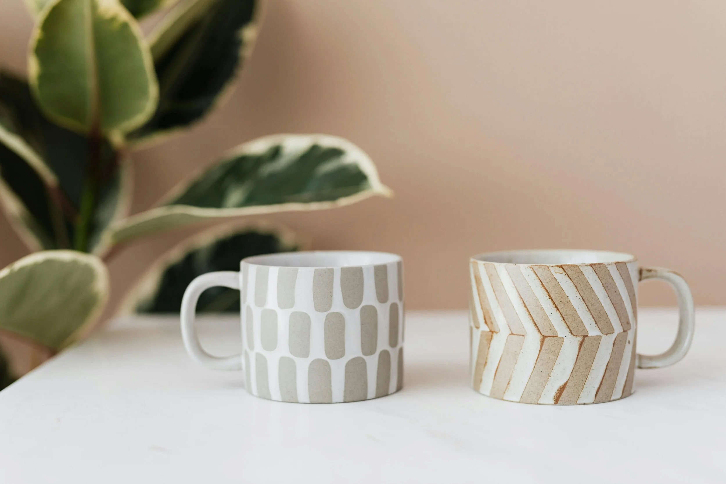 Deux tasses en céramique, une avec un motif de rayures blanches et beiges, l'autre avec un motif de lignes verticales grises et blanches, sur une surface blanche avec une plante verte en arrière-plan.