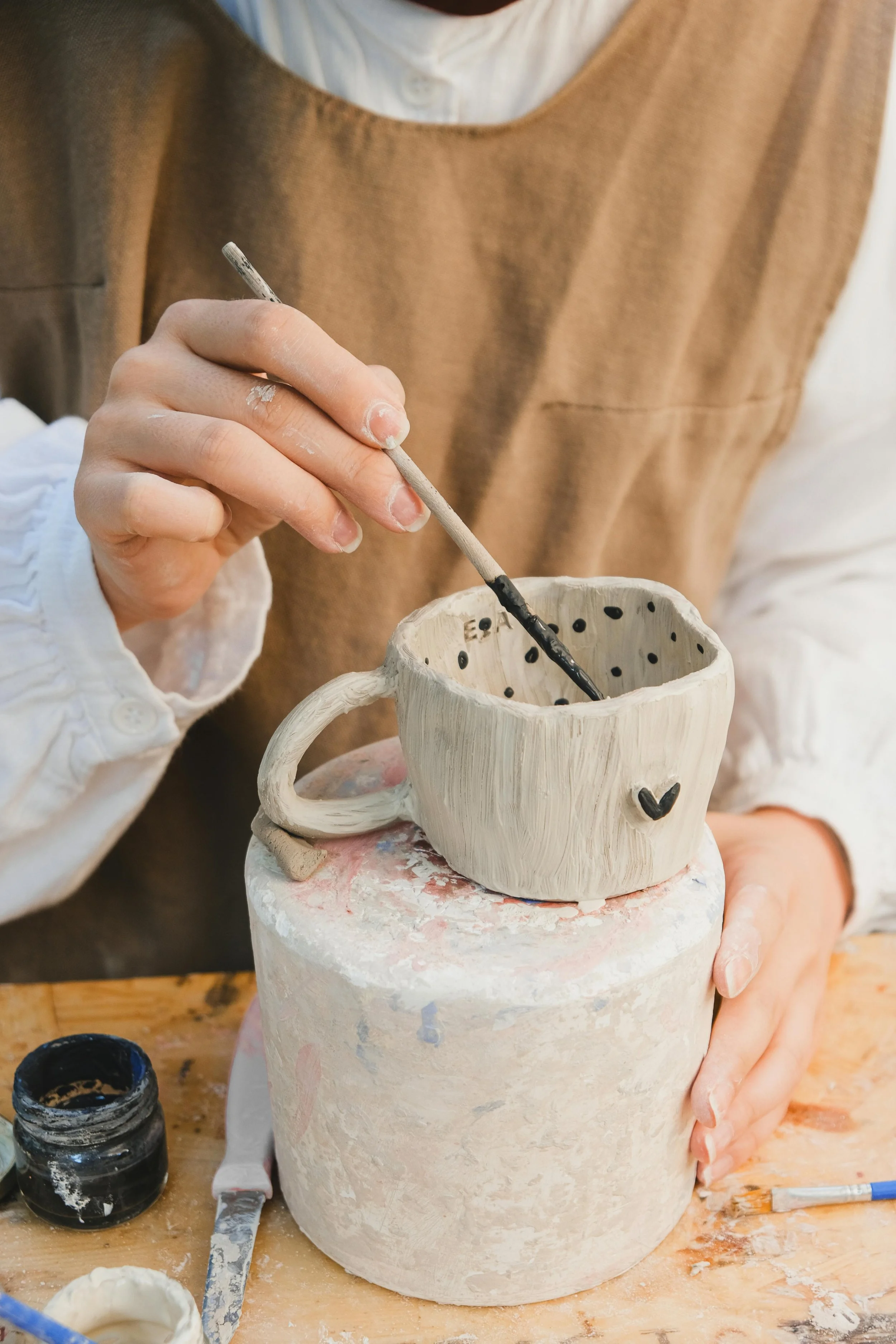 Une personne peint un mug en céramique blanc avec une peinture noire, créant un dessin de cœur sur le mug, dans un atelier de poterie.