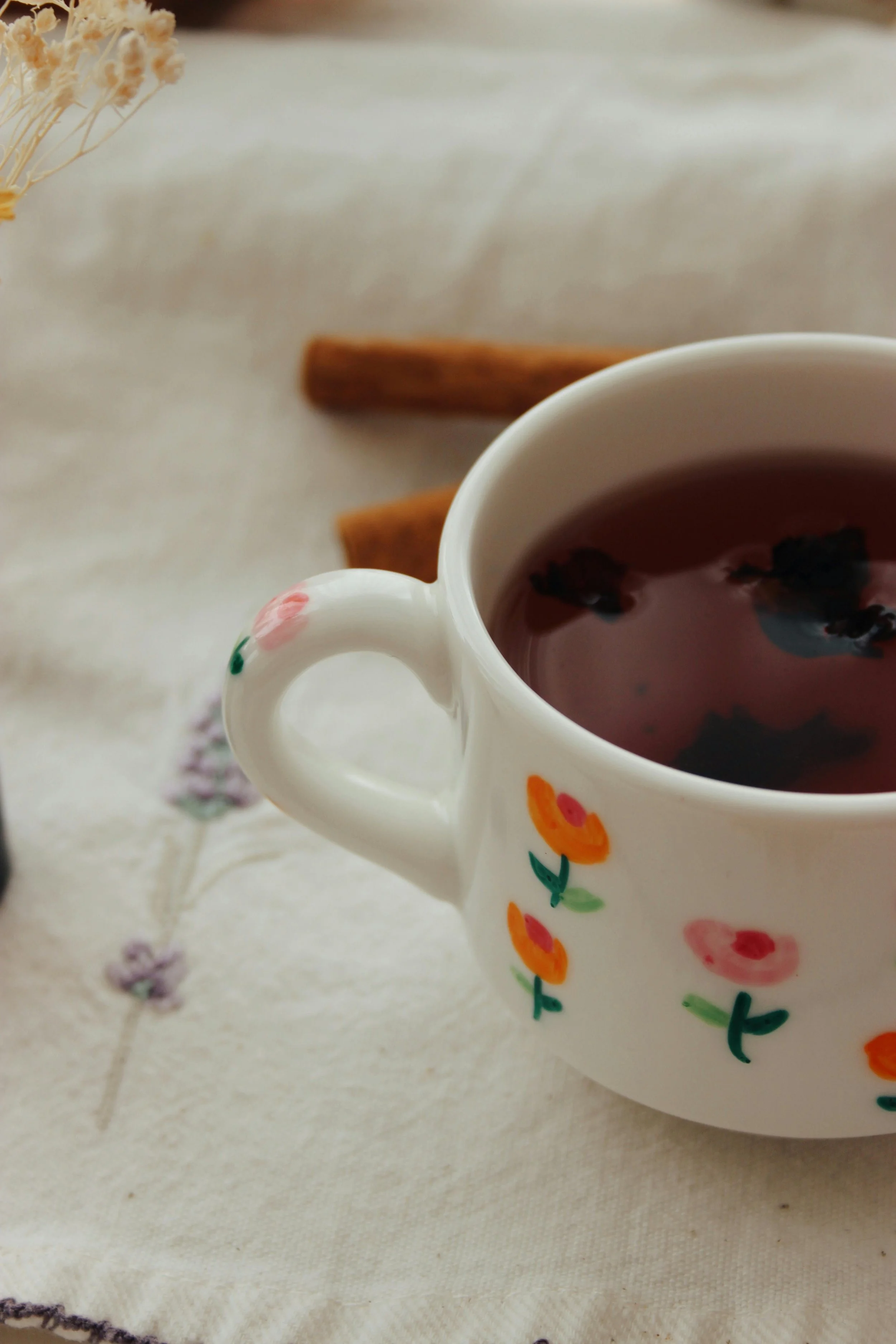Une tasse de thé ou de tisane sur une nappe blanche à motifs floraux, avec deux bâtons de cannelle à l’arrière et une branche de fleurs séchées à gauche.