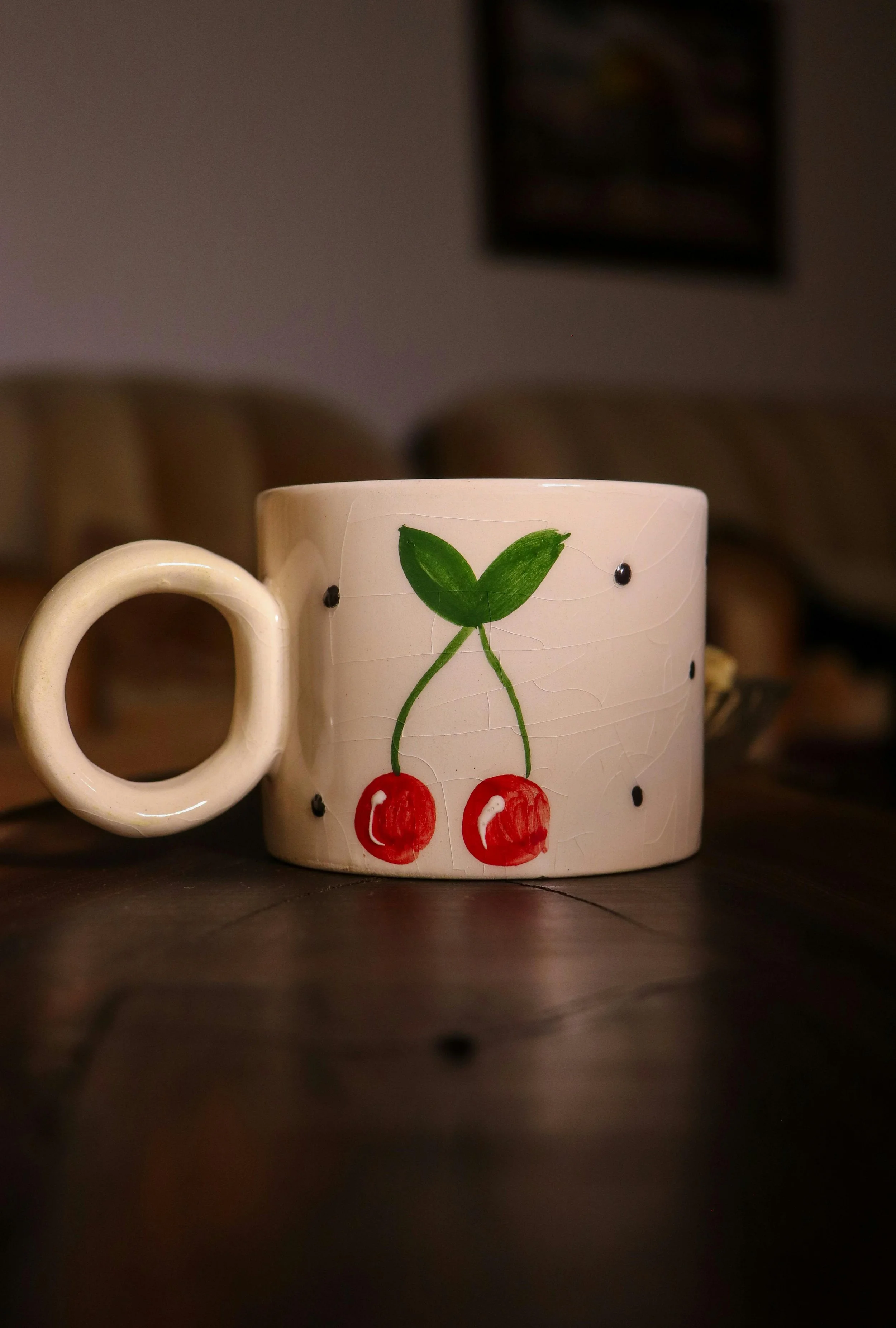 Tasse en céramique blanche avec décoration de cerises rouges et feuillage vert, placée sur une surface en bois, fond flou et légèrement sombre.