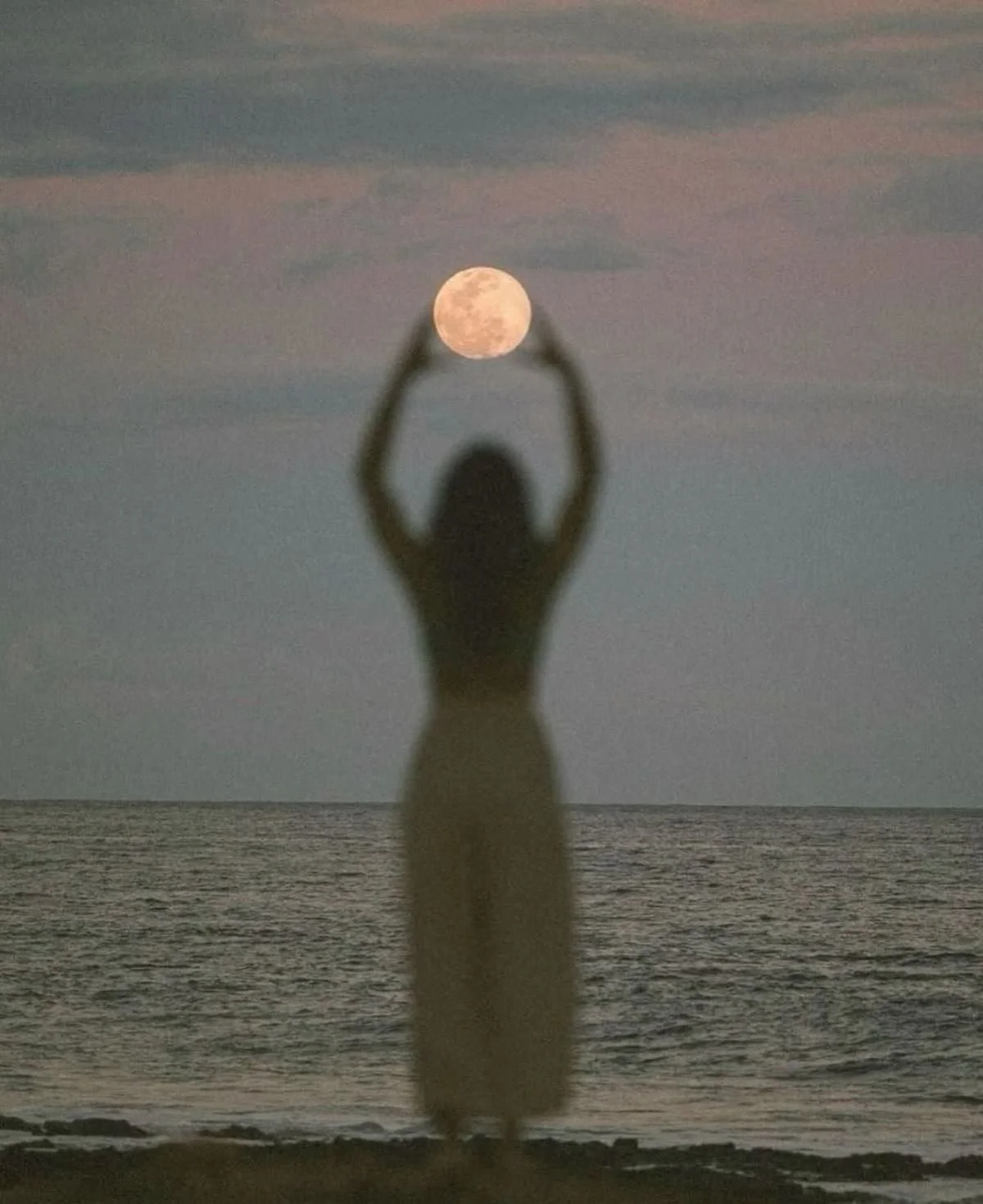 A woman standing on the beach with arms raised, forming a frame around the full moon in the sky during sunset.