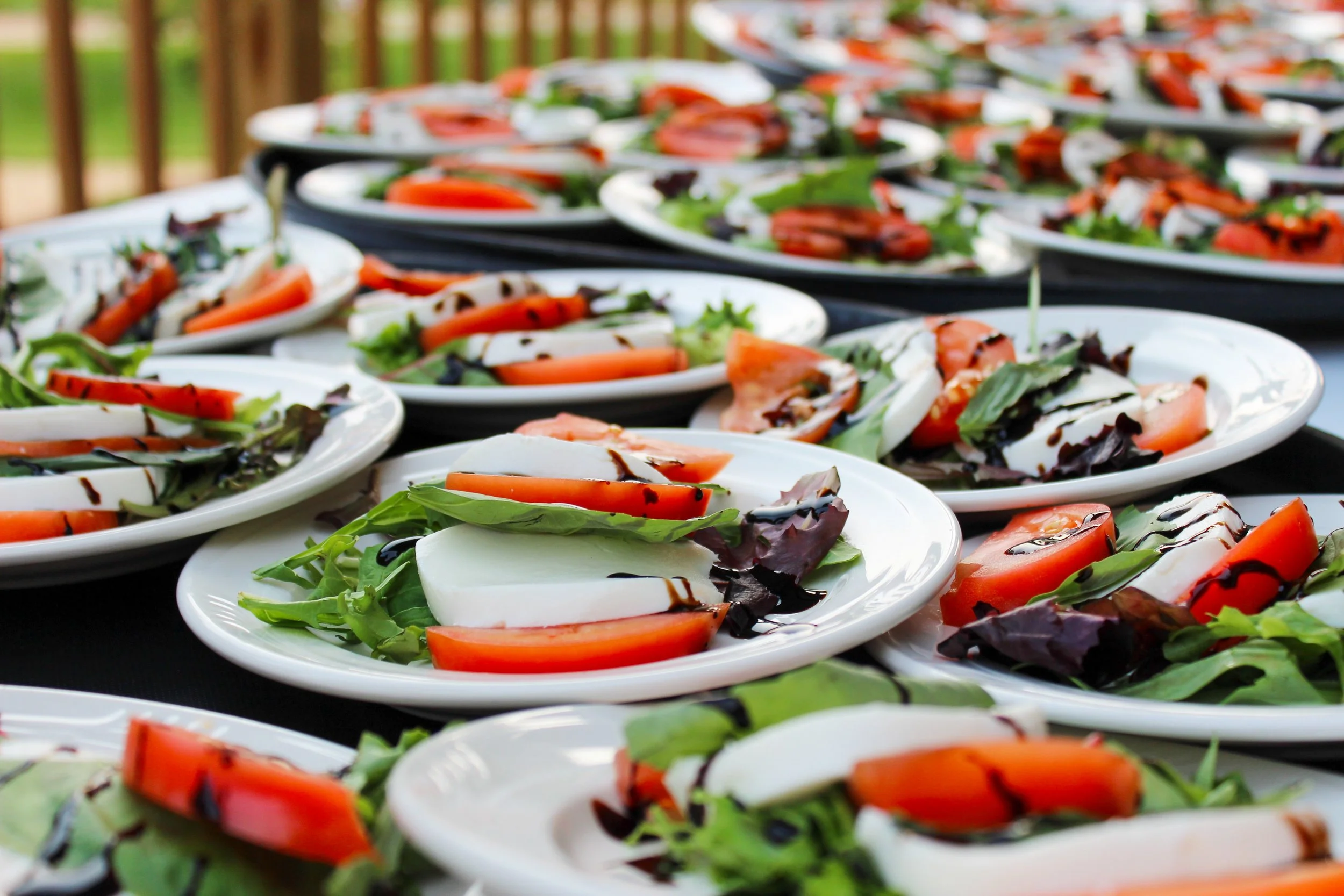 Multiple plates of caprese salad with sliced tomatoes, mozzarella cheese, basil, and balsamic glaze, arranged on a table outdoors.