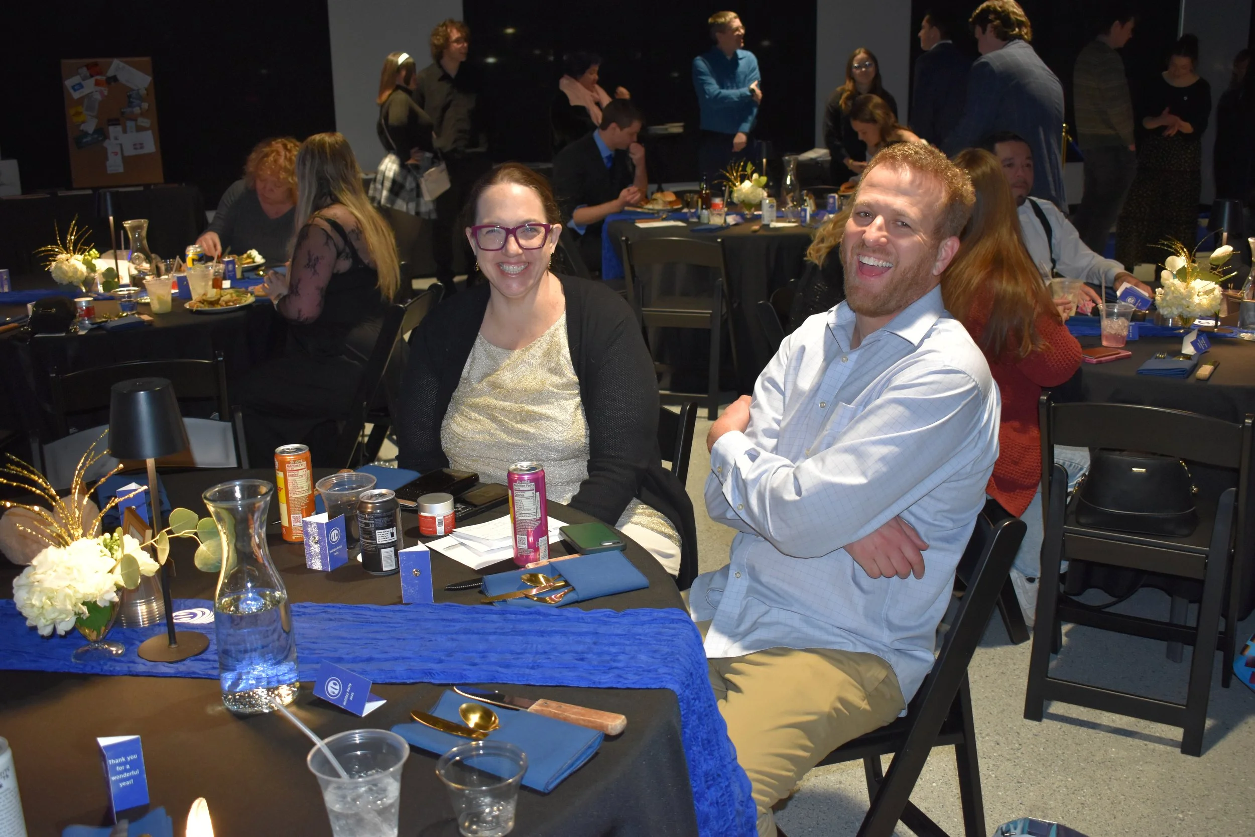 Group of people at a banquet, two people smiling at the camera in the foreground, decorated tables with flowers, drinks, and place cards in the background.