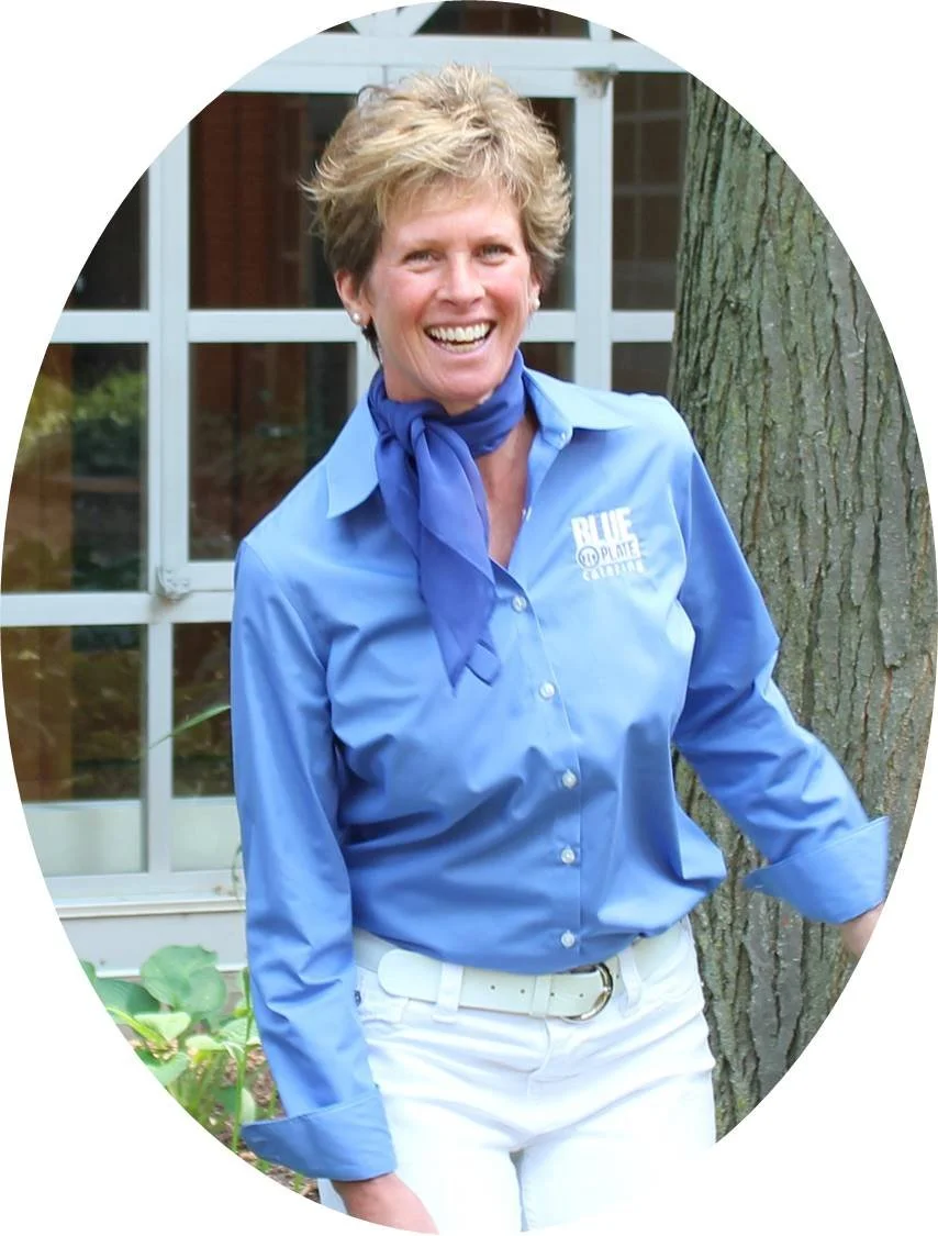 A smiling woman with short, light-colored hair wearing a blue button-up shirt with a logo and a blue neckerchief, standing outdoors next to a tree and a large windowed building.