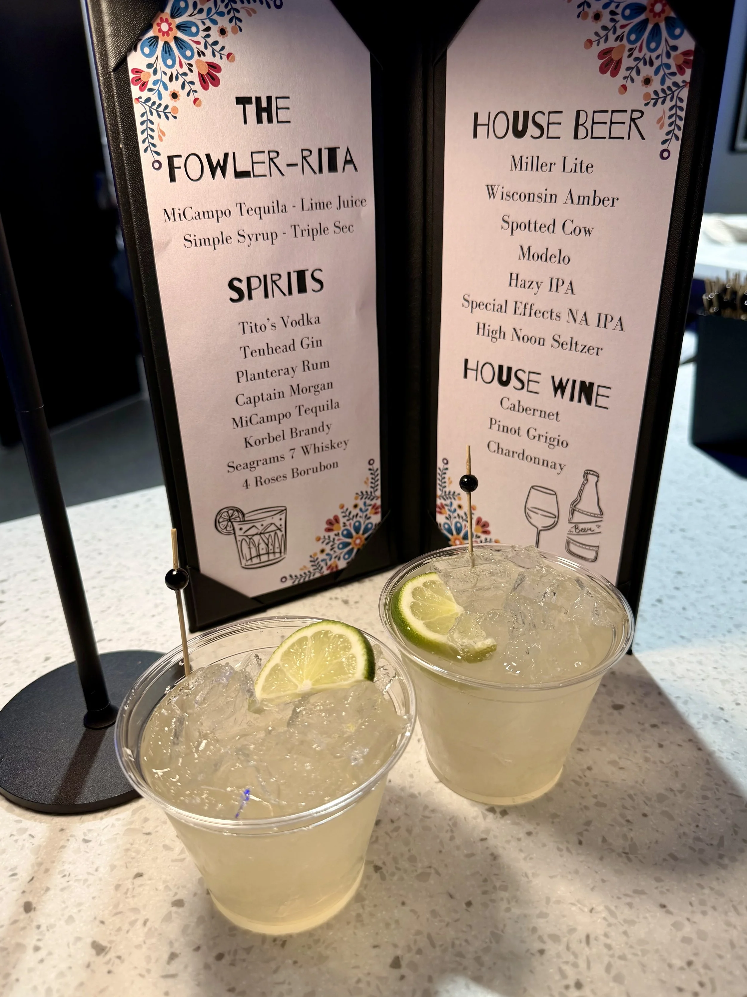 Two cocktails with lemon wedges served in clear plastic cups and a black stand with a menu. The menu lists drinks, spirits, house beer, and house wine, decorated with floral patterns.