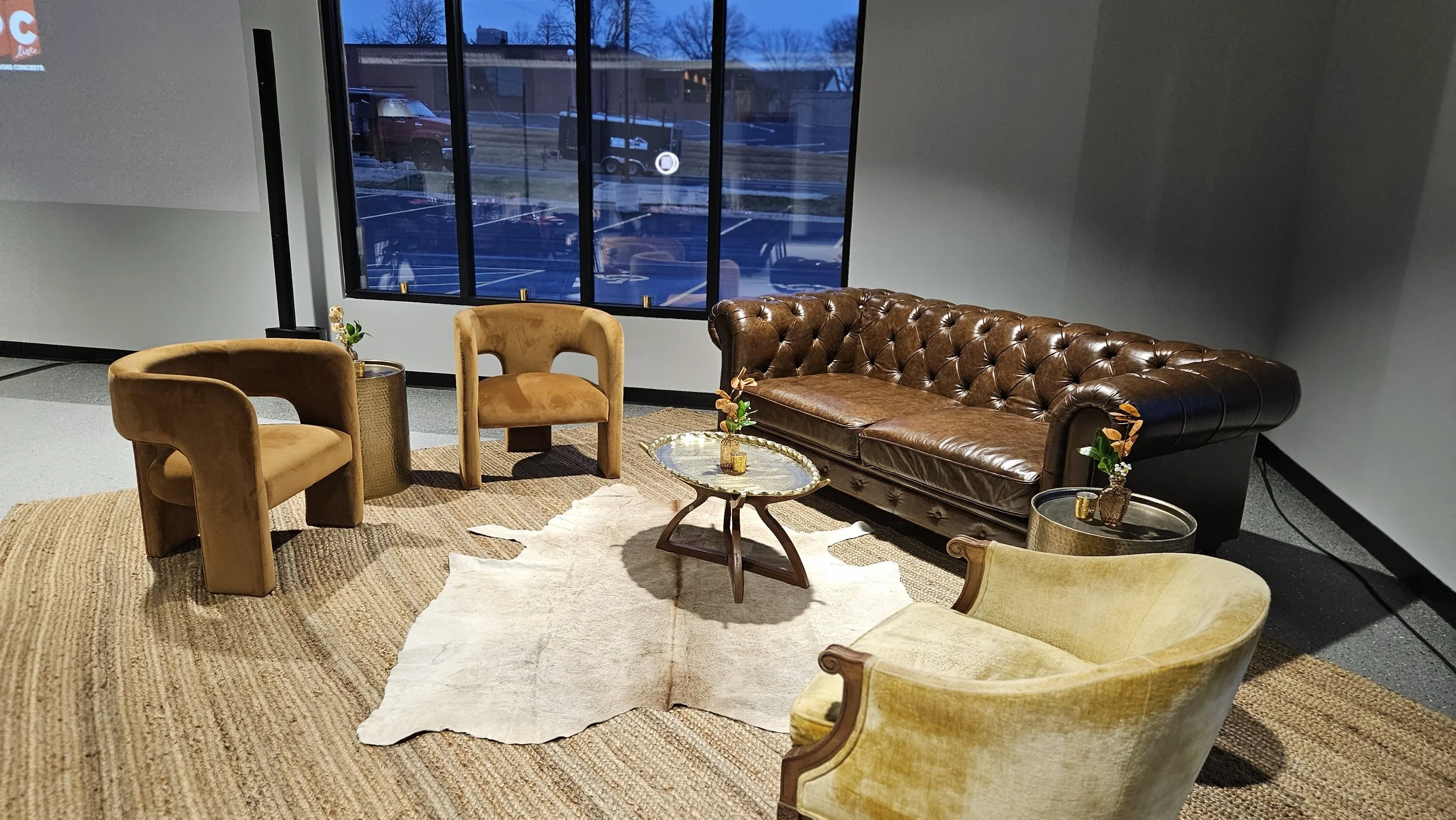 Cozy waiting area with a brown leather tufted sofa, two mustard-colored velvet armchairs, a small round coffee table with a decorative tray, and a white rug on a brown woven mat in front of a large window showing a parking lot and cars outside.