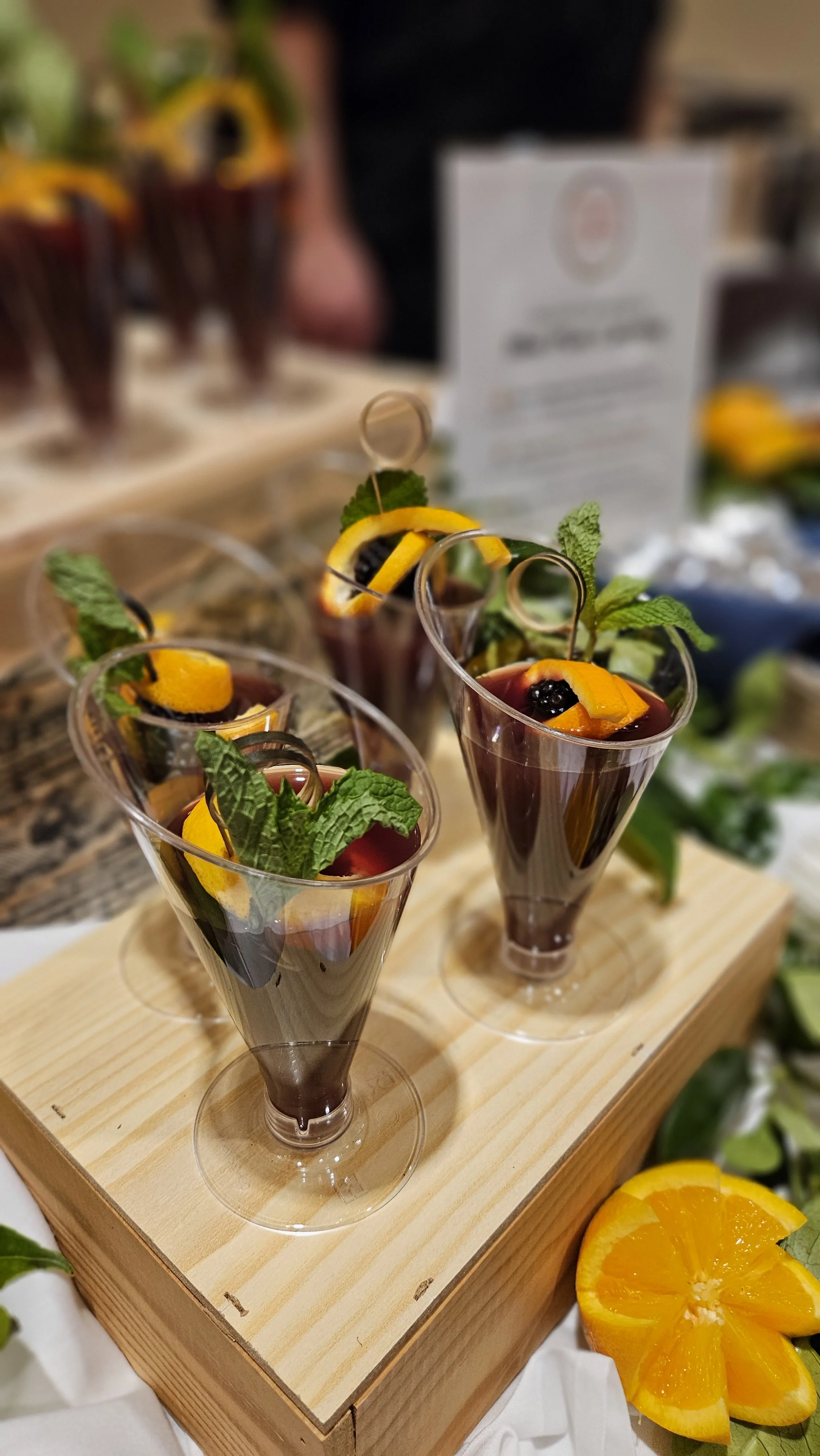 Three glasses of red wine or sangria garnished with orange slices, blueberries, mint leaves, and skewers, presented on a wooden board.