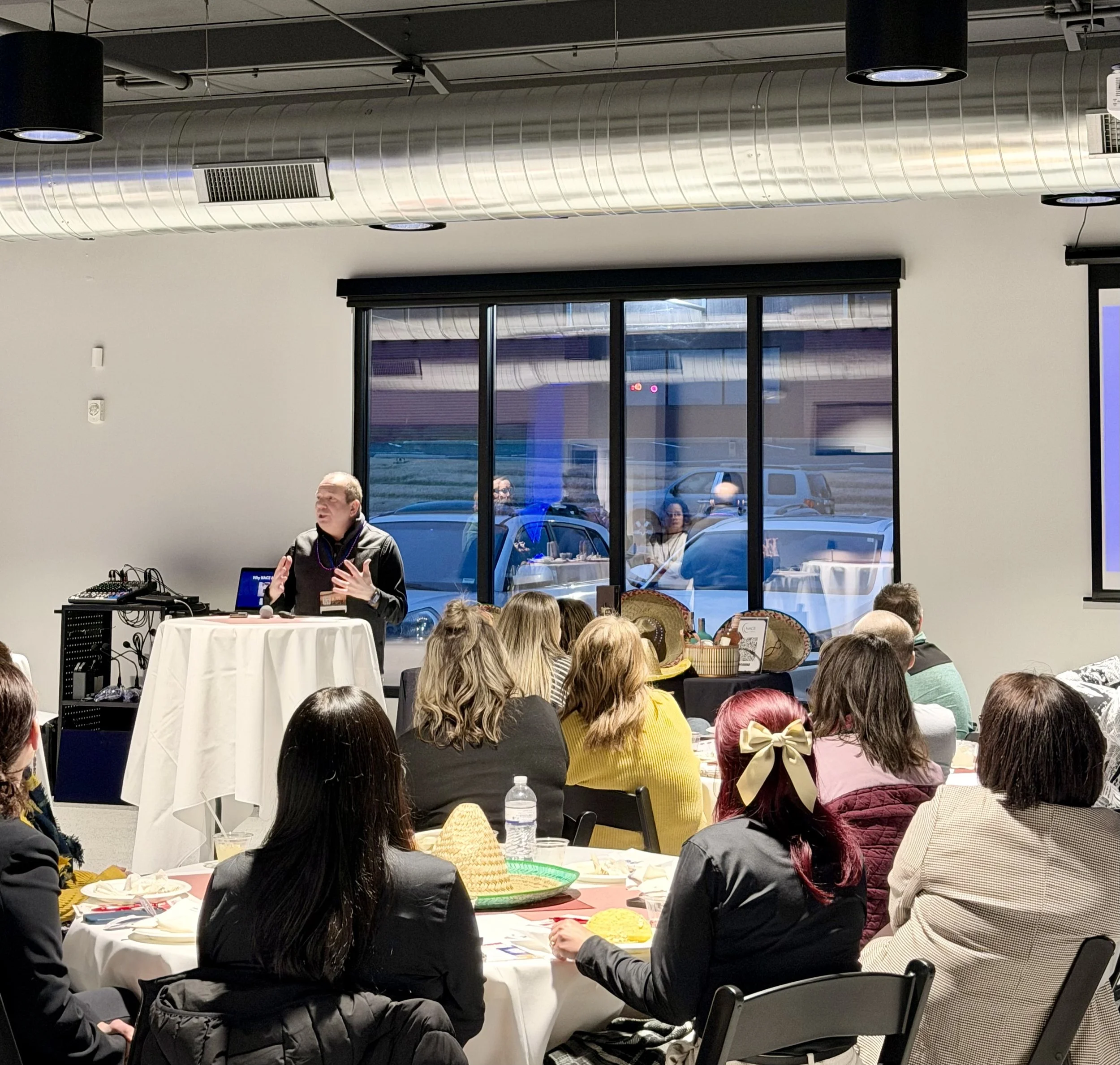 A man giving a presentation to a group of people seated at round tables in a modern conference room. There is a large window behind him showing parked cars outside, and the room has ceiling vents and black pendant lights.
