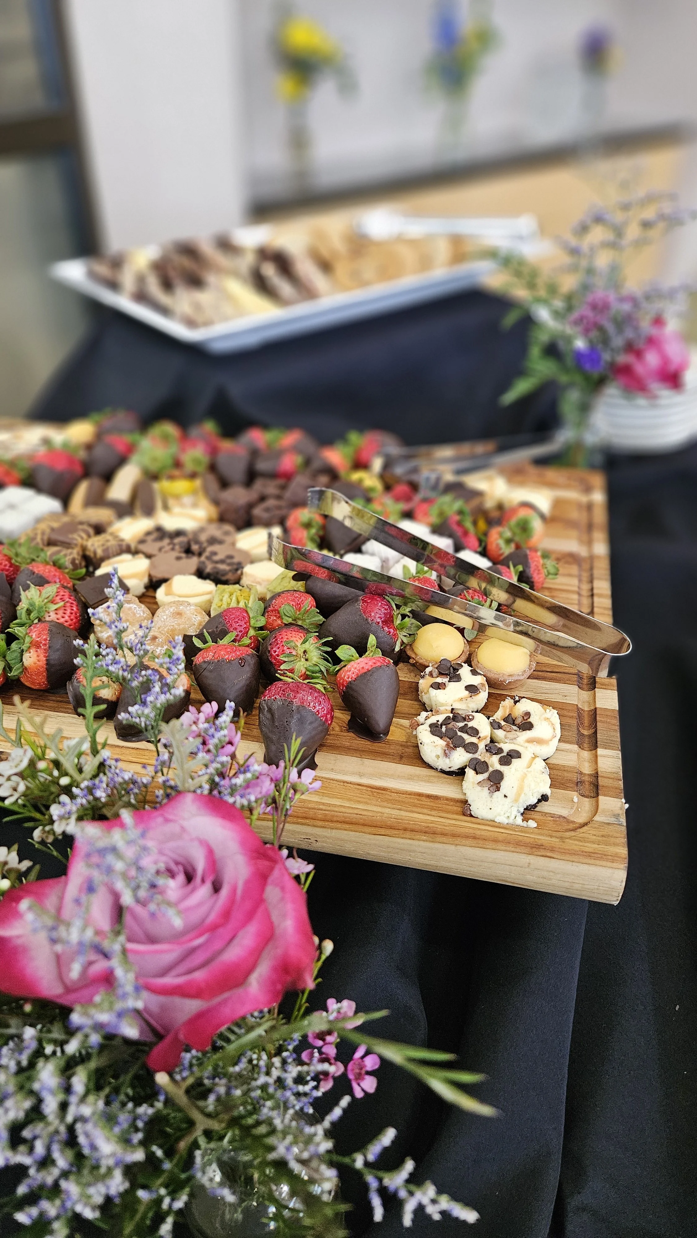 A wooden tray with assorted candies, chocolates, and strawberries, surrounded by colorful flowers on a black tablecloth.