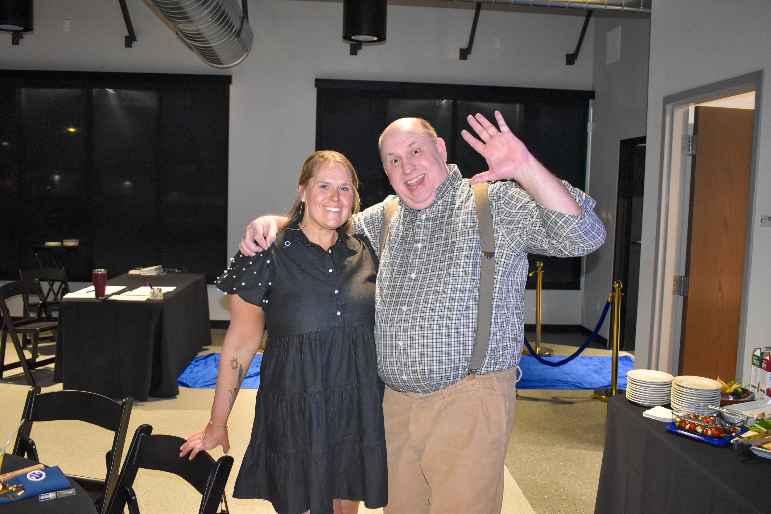 A woman and a man standing indoors, smiling, with the woman’s arm around the man’s shoulder and the man waving at the camera. The woman is wearing a black dress with pearl embellishments on the sleeves, and the man is in a plaid shirt with suspenders.
