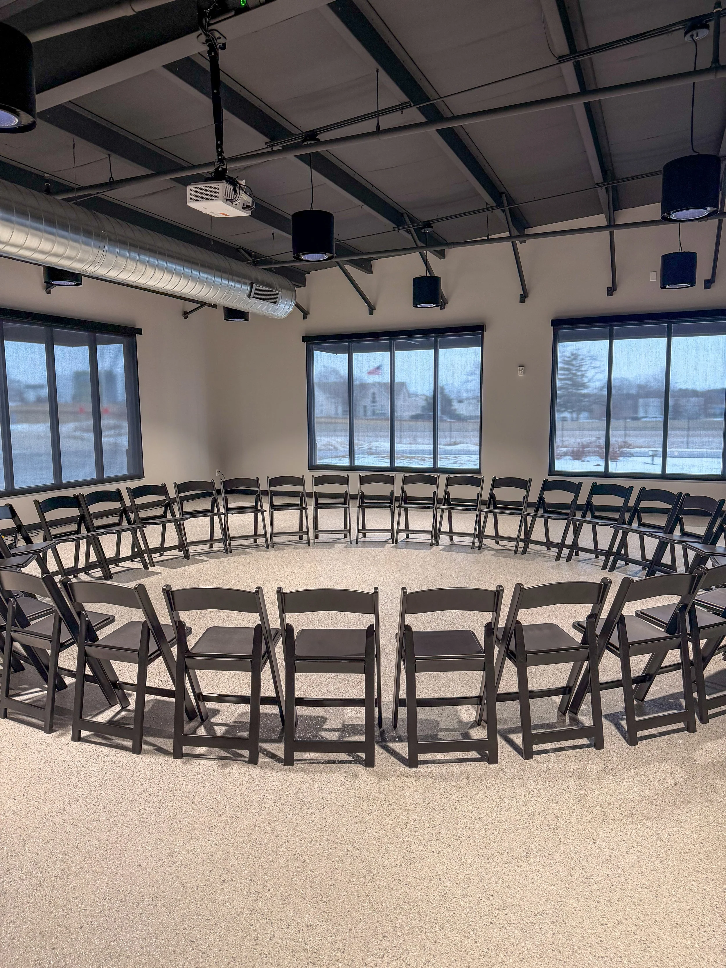 Empty room with chairs arranged in a large circle, windows with shades, and ceiling with exposed ductwork and black hanging lights.