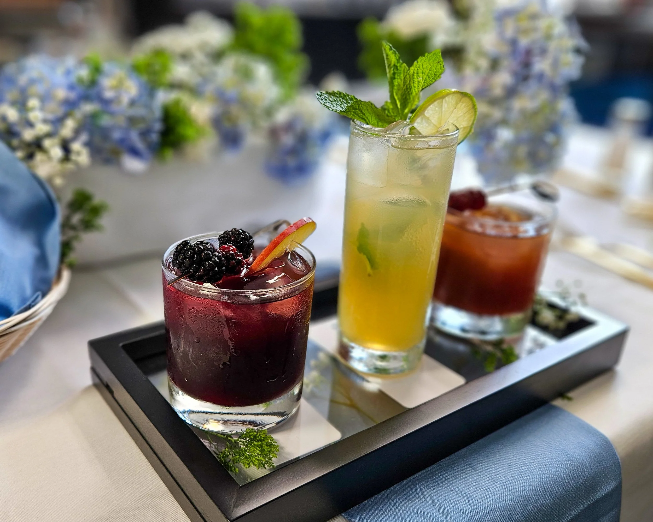 Three colorful cocktails on a tray, garnished with fresh fruit and herbs, set on a table with floral decorations in the background.