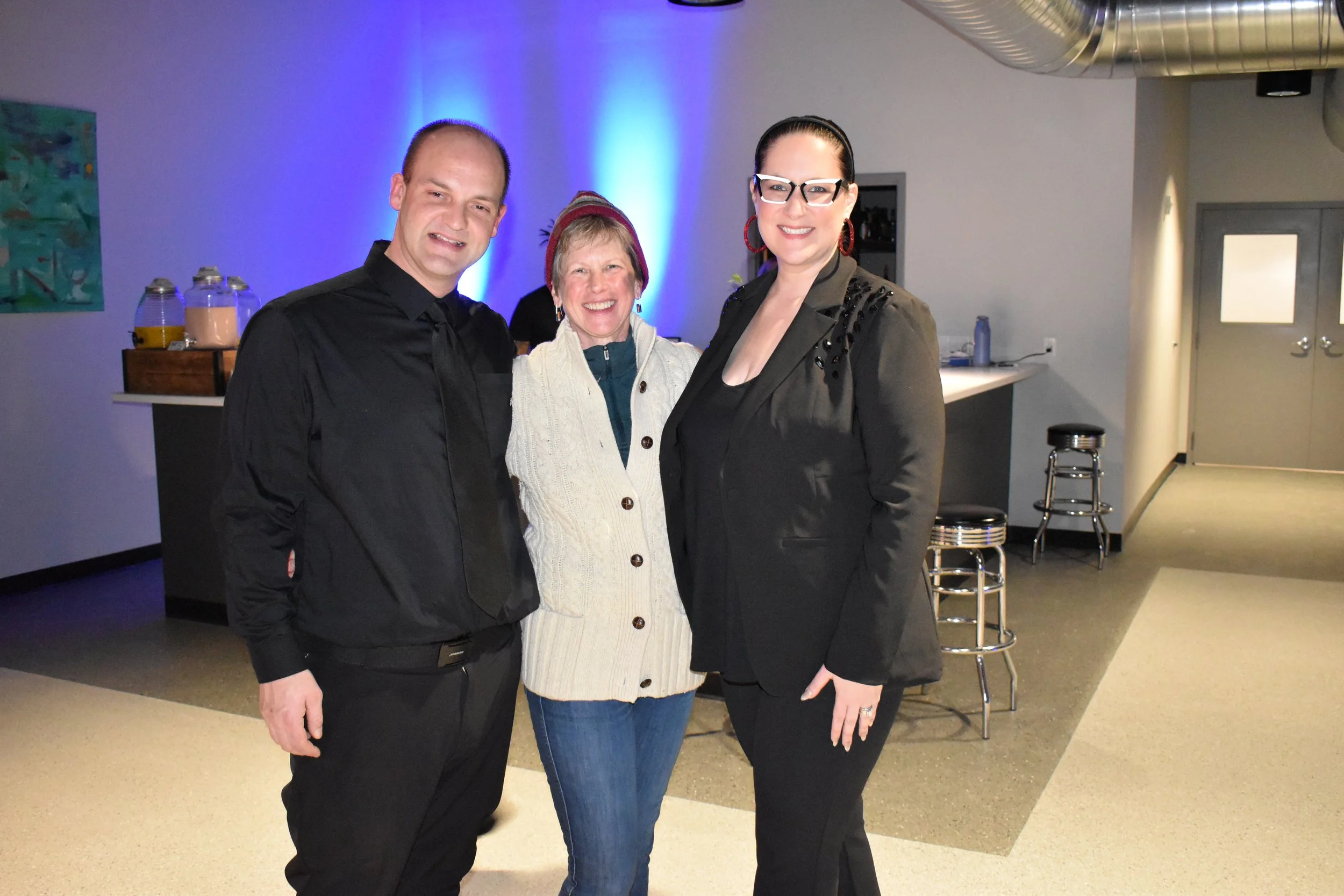 Three people smiling and standing close together in an indoor setting with modern decor, including bar stools and a counter in the background.