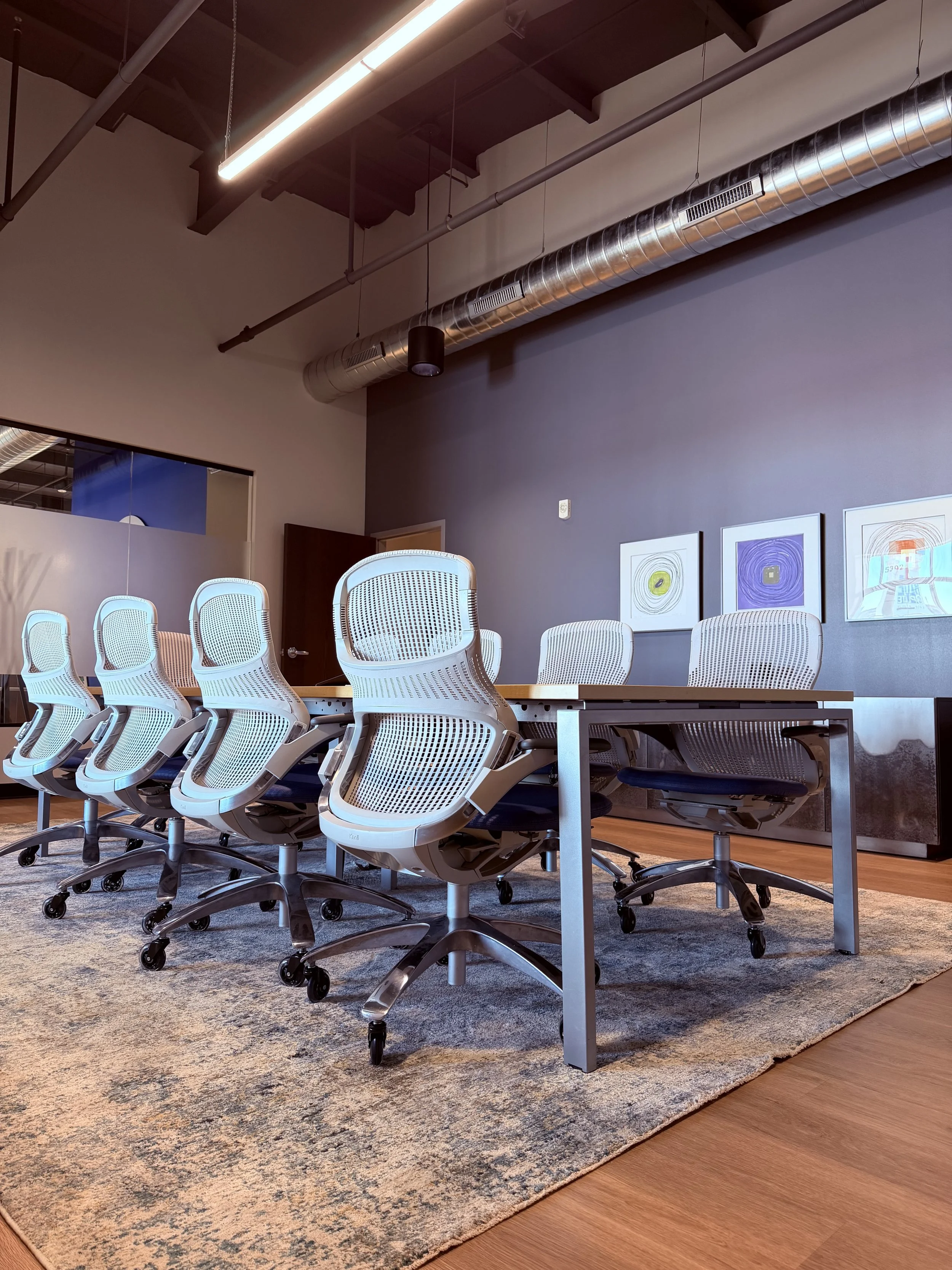 Modern conference room with six white ergonomic chairs with mesh backs and armrests, set around a rectangular wooden table. The room features a dark accent wall with three framed abstract art pieces and contemporary lighting fixtures from an industri
