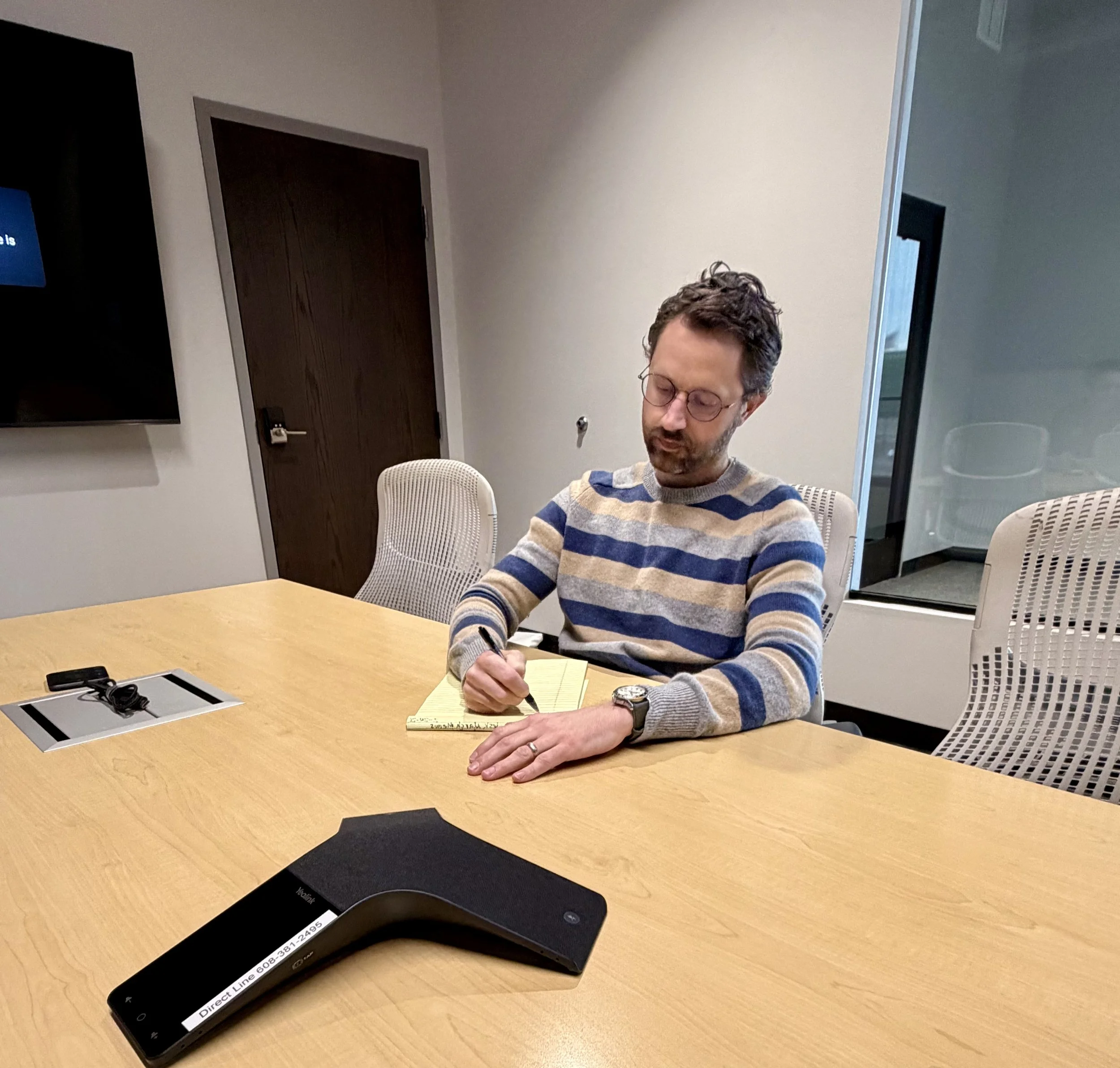 A man with glasses and a beard, wearing a striped sweater, sits at a conference table taking notes in a yellow notebook during a meeting or interview in a modern office.