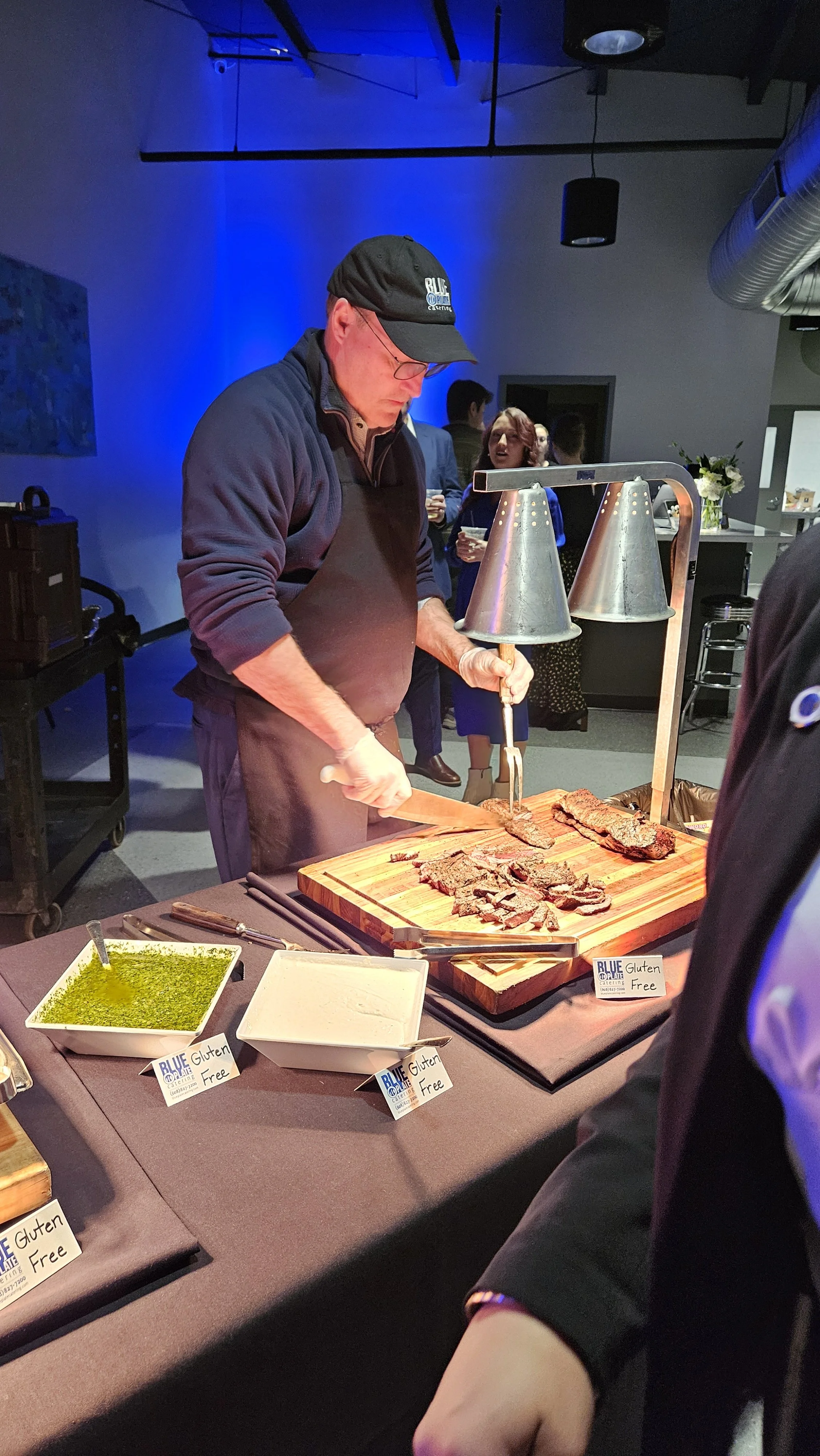 A man slicing cooked beef at a buffet table, with bowls of green sauce labeled gluten-free nearby.