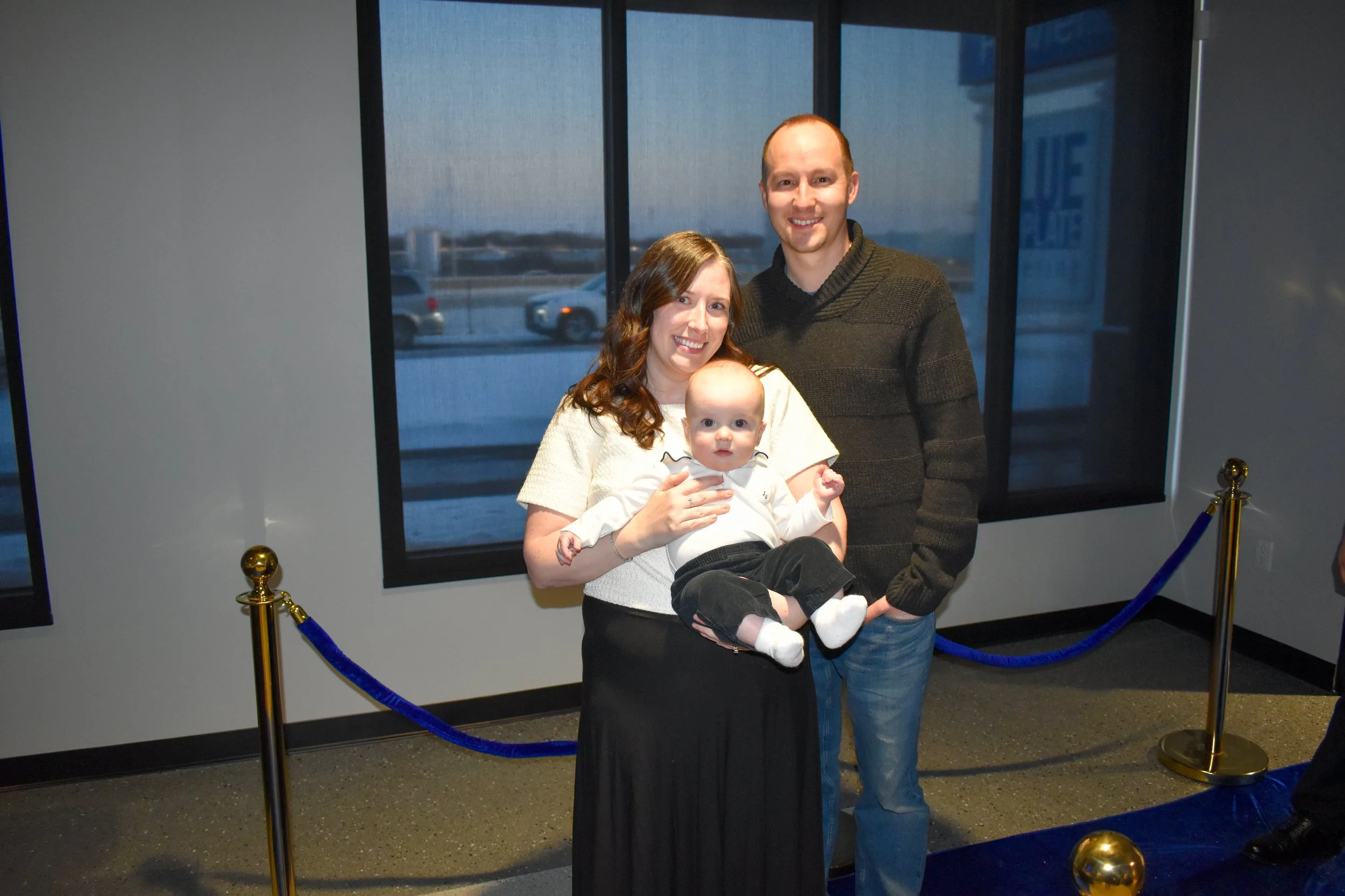 Family of three posing at an indoor event, with a woman holding a baby boy and a man standing beside them, behind a velvet rope barrier.