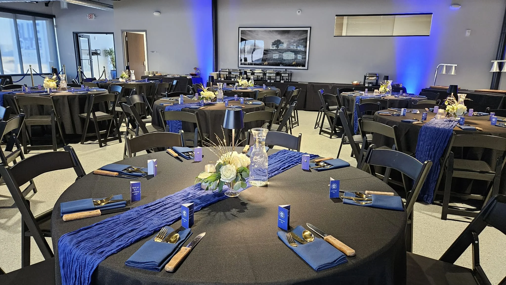 Event banquet hall with round tables decorated with blue table runners, white floral centerpieces, and place settings with gold utensils and blue napkins. The room has large windows, a framed black-and-white landscape photograph on the wall, and blue uplighting on some walls.