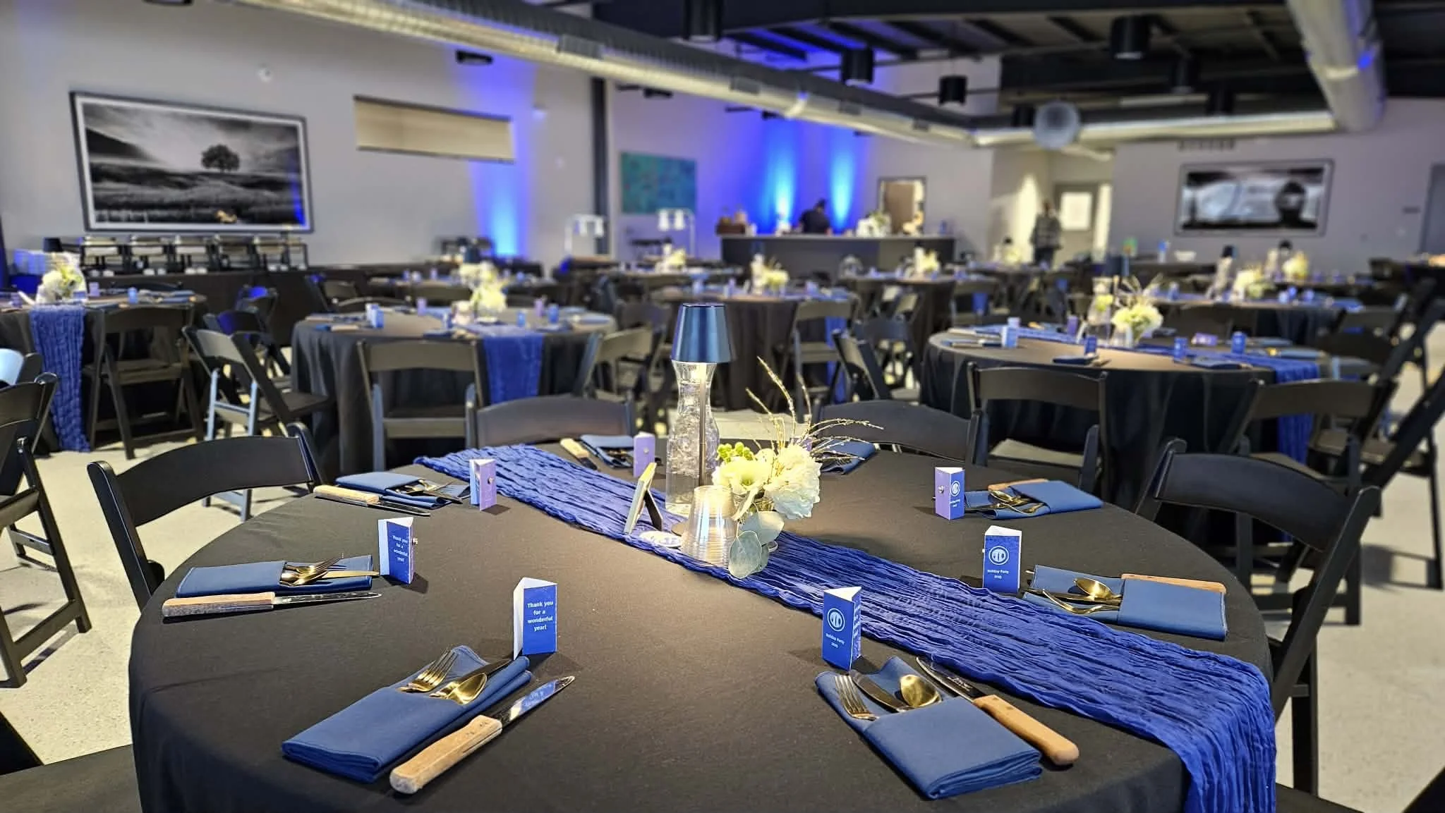 A banquet hall decorated for an event with round tables covered in black tablecloths and blue napkins, featuring centerpieces with white flowers, a blue table runner, and a tall glass vase. The hall has blue uplighting, framed pictures on the walls, 