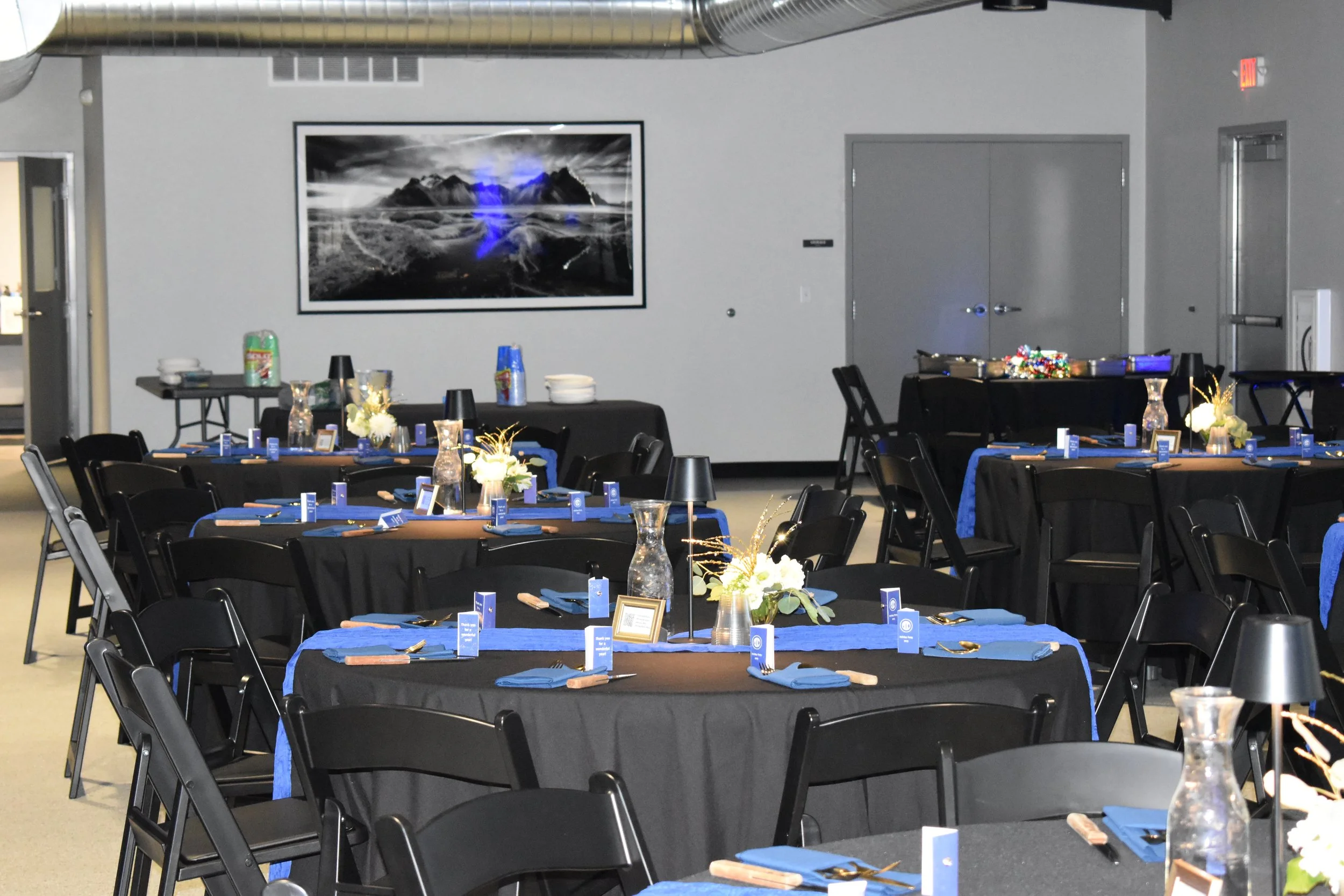 Indoor event space with round tables covered in black and blue tablecloths, decorated with vases of white flowers, small lamps, and place settings, ready for a celebration or banquet.