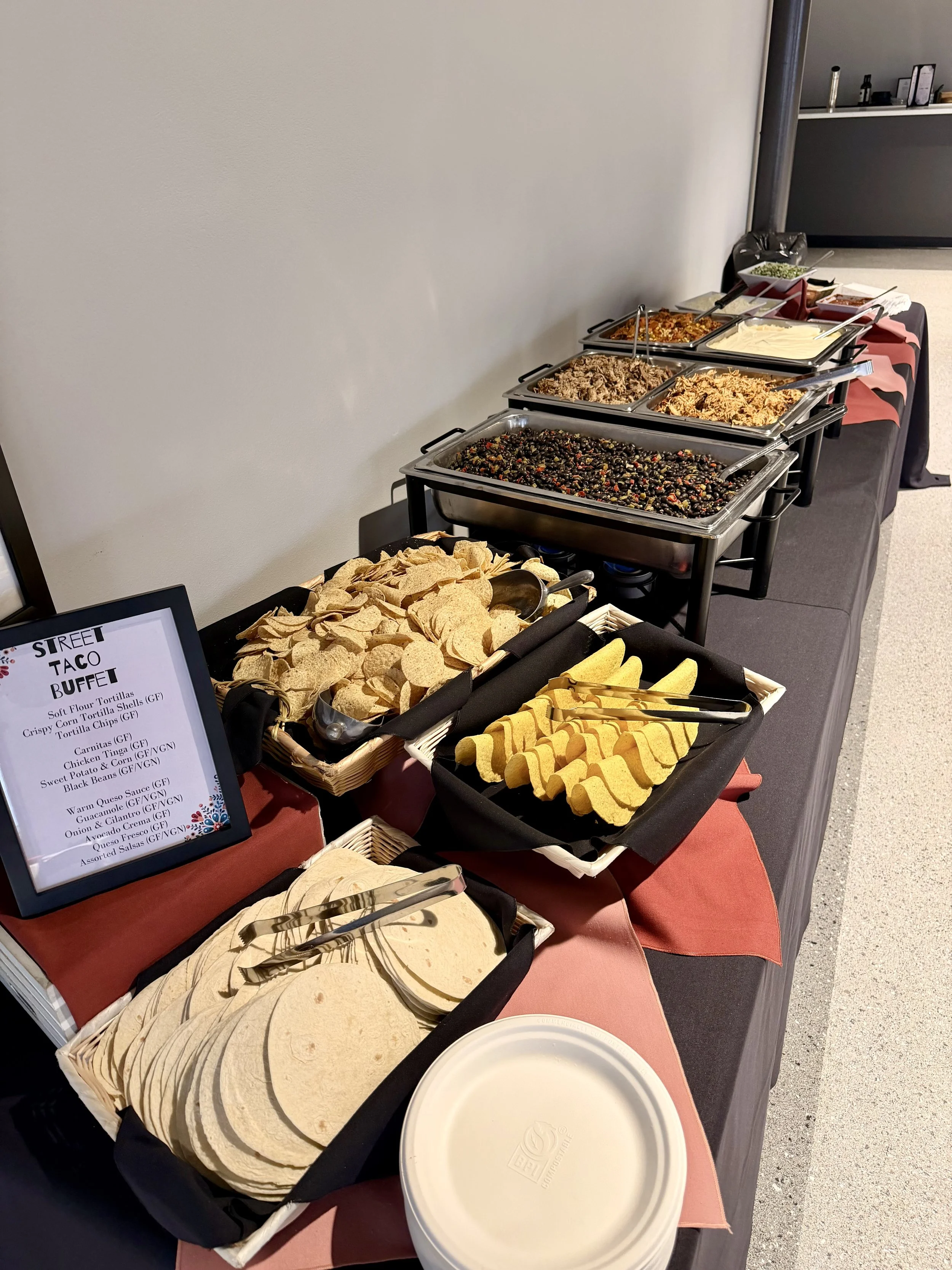 Buffet table with various taco toppings, including shredded meat, beans, cheese, and chips, with a sign indicating 'Street Taco Buffet'.